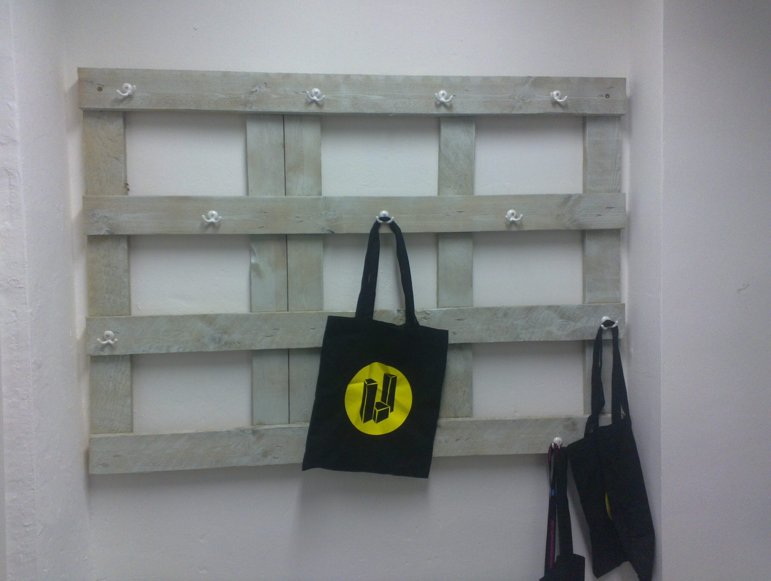 Holzpalette an Wand montiert mit Metallhaken, daran hängen schwarze Taschen.