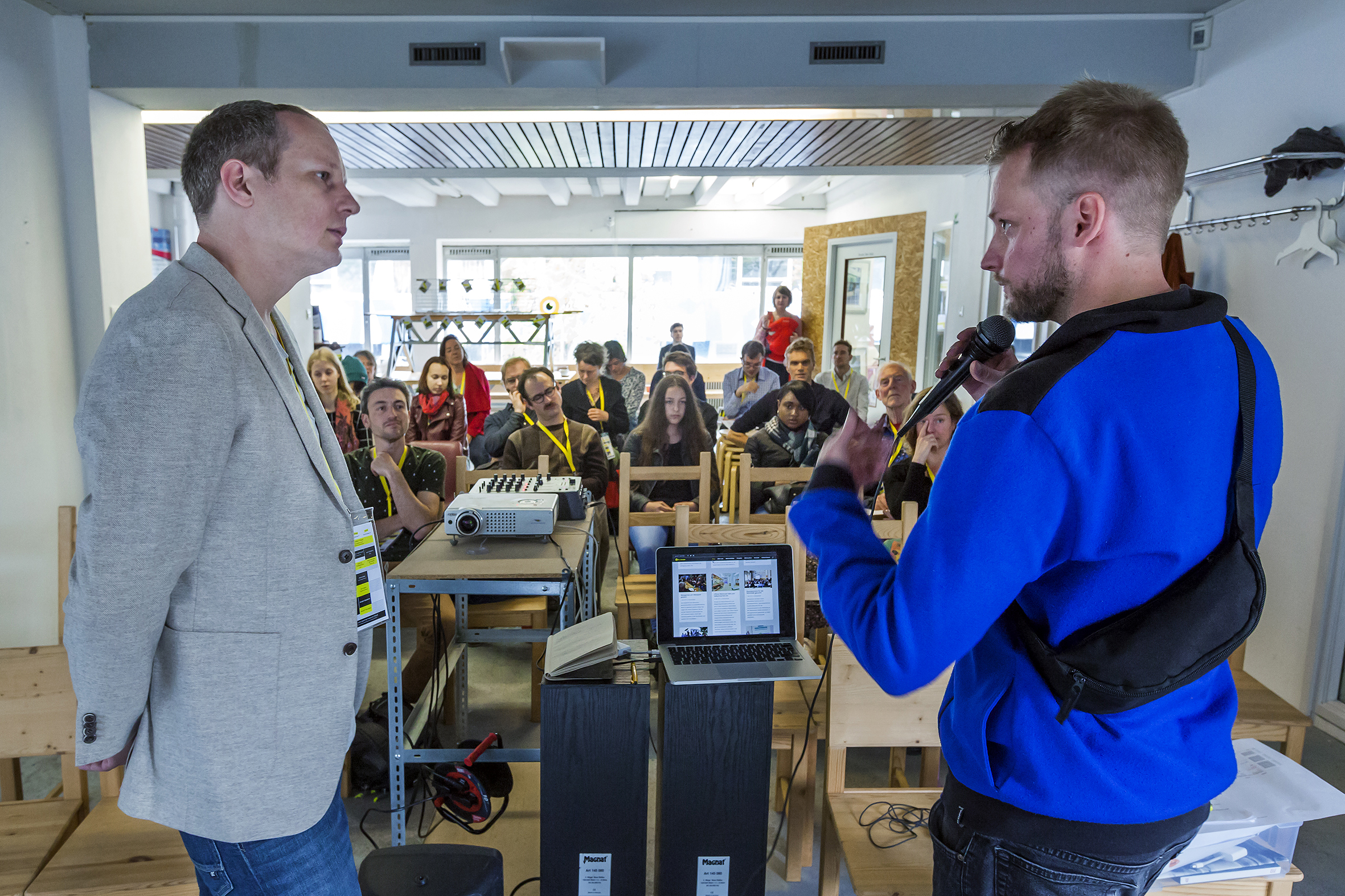 Zwei Männer präsentieren vor Publikum mit Mikrofon an Tischen mit technischer Ausrüstung.