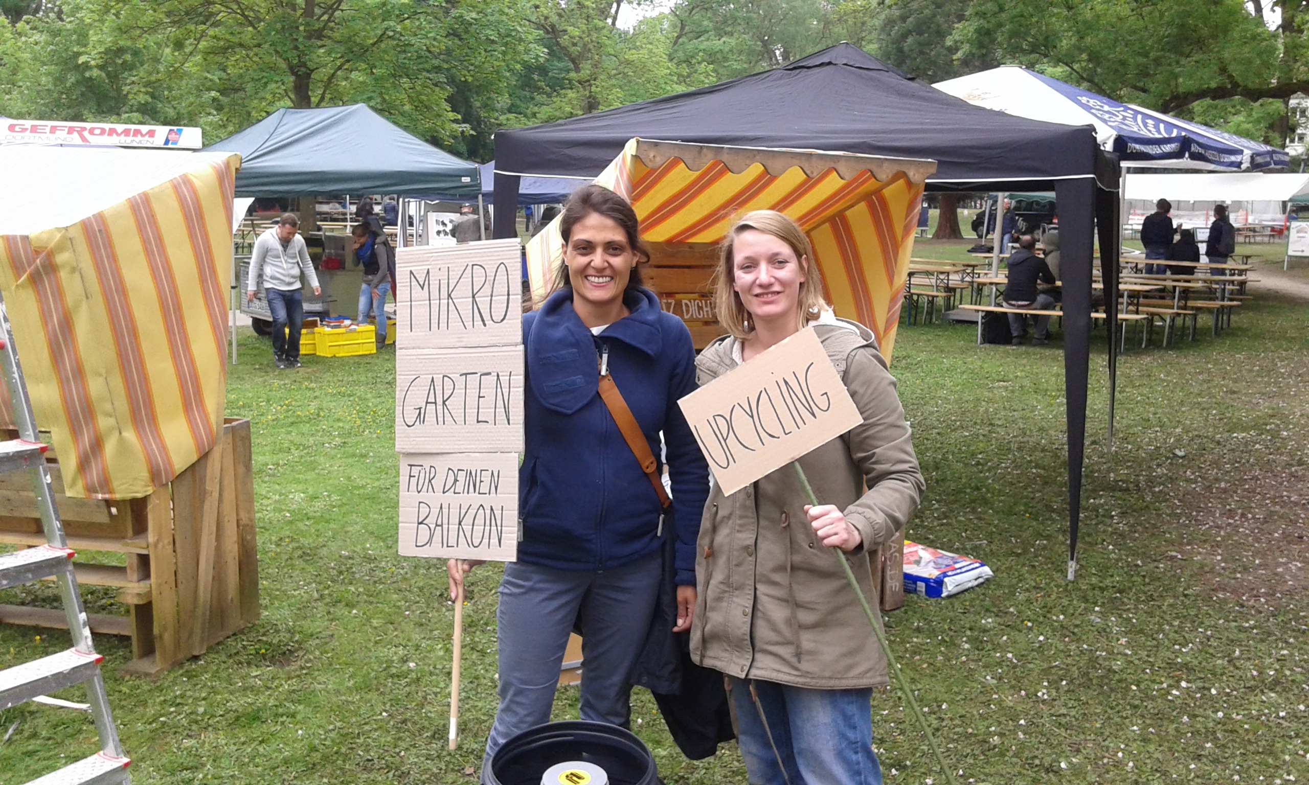 Zwei Frauen präsentieren bei einem Straßenfest Schilder zu Mikrogärten und Balkonbegrünung unter schwarzem Zelt.