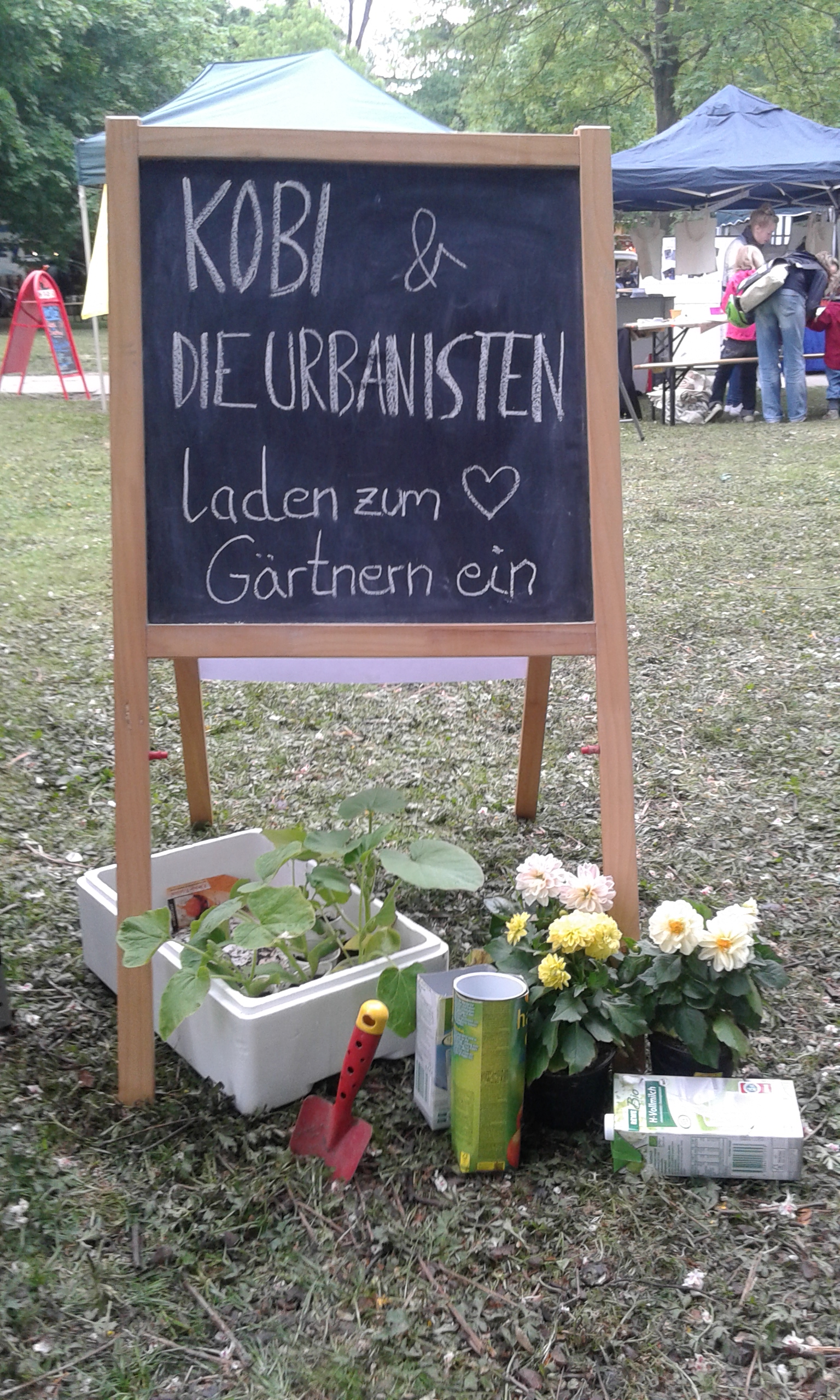 Schautafel mit Aufschrift Kobi & Die Urbanisten auf Marktplatz mit Blumenkübeln.