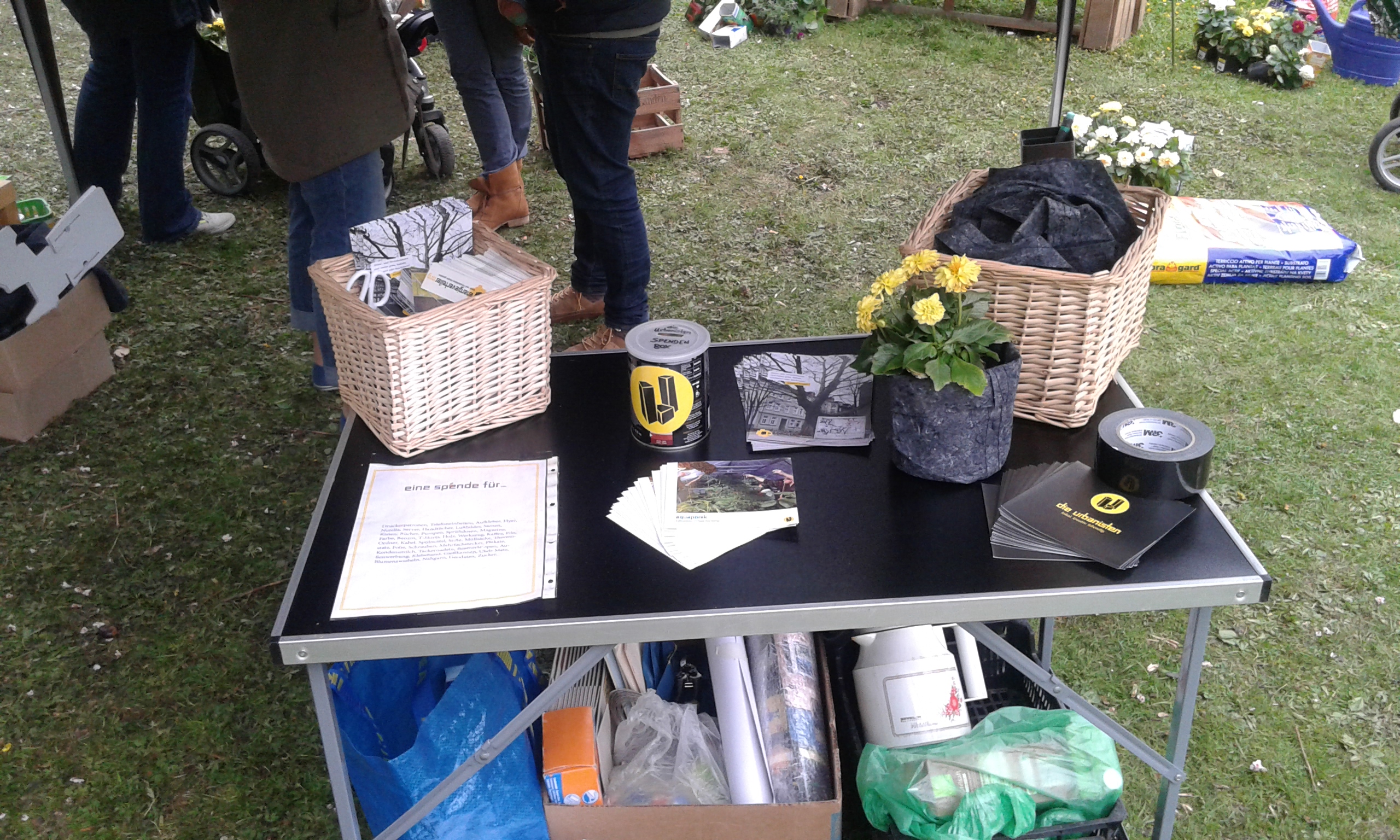 Informationsstand mit Körben, Broschüren und Blumen auf Grasfläche bei Veranstaltung.