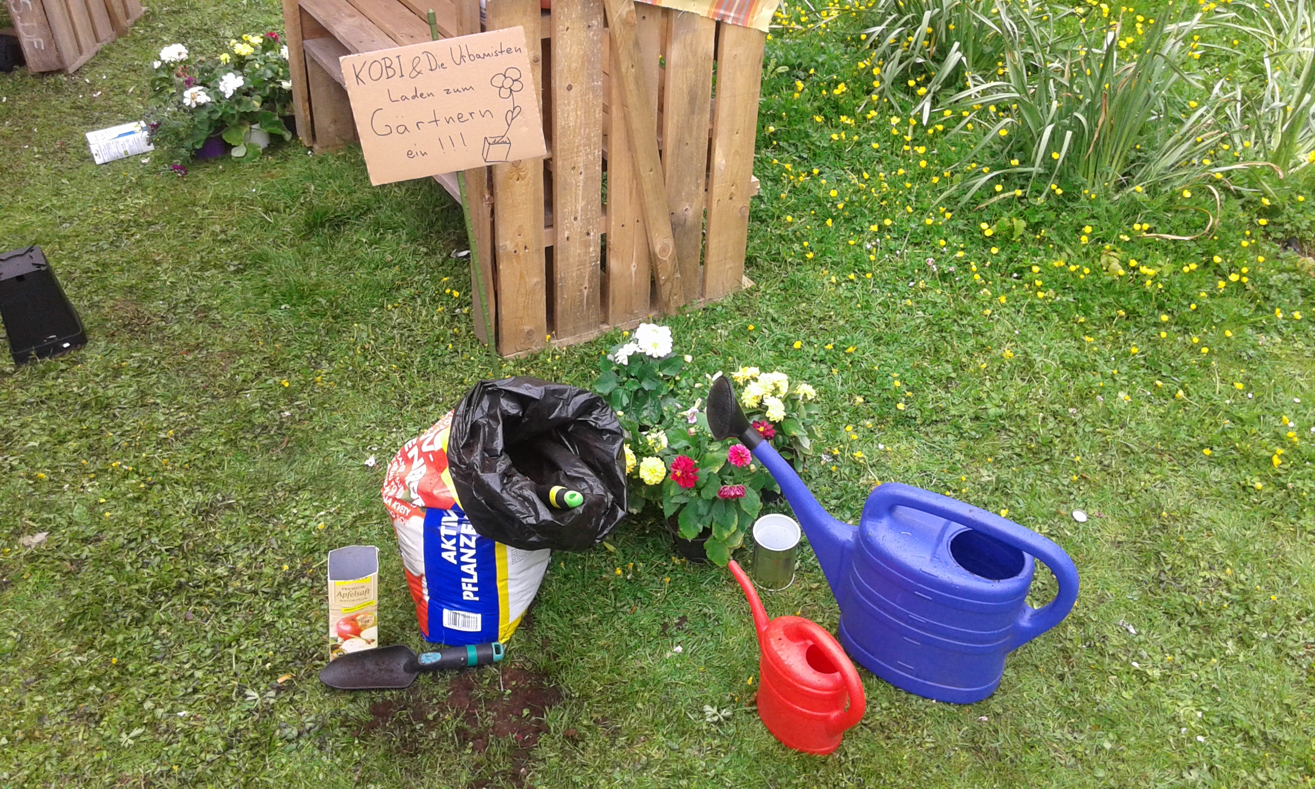 Gemeinschaftsgarten mit Holzpaletten-Hochbeet, Blumen, Gießkanne und Hinweisschild.
