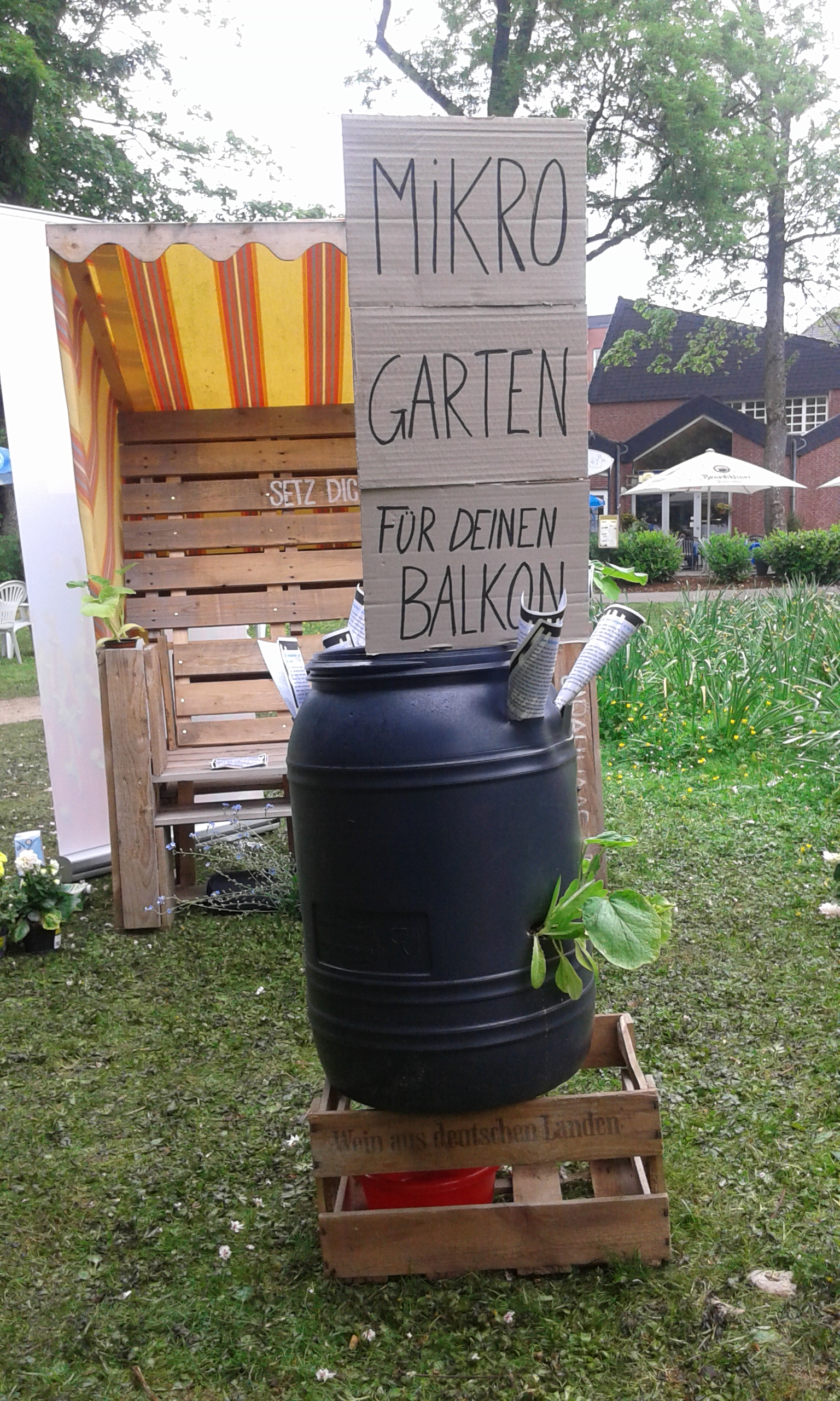Selbstgebaute Mikrogarten-Installation aus Holzpaletten, schwarzem Behälter und Beschriftung in einem Garten.