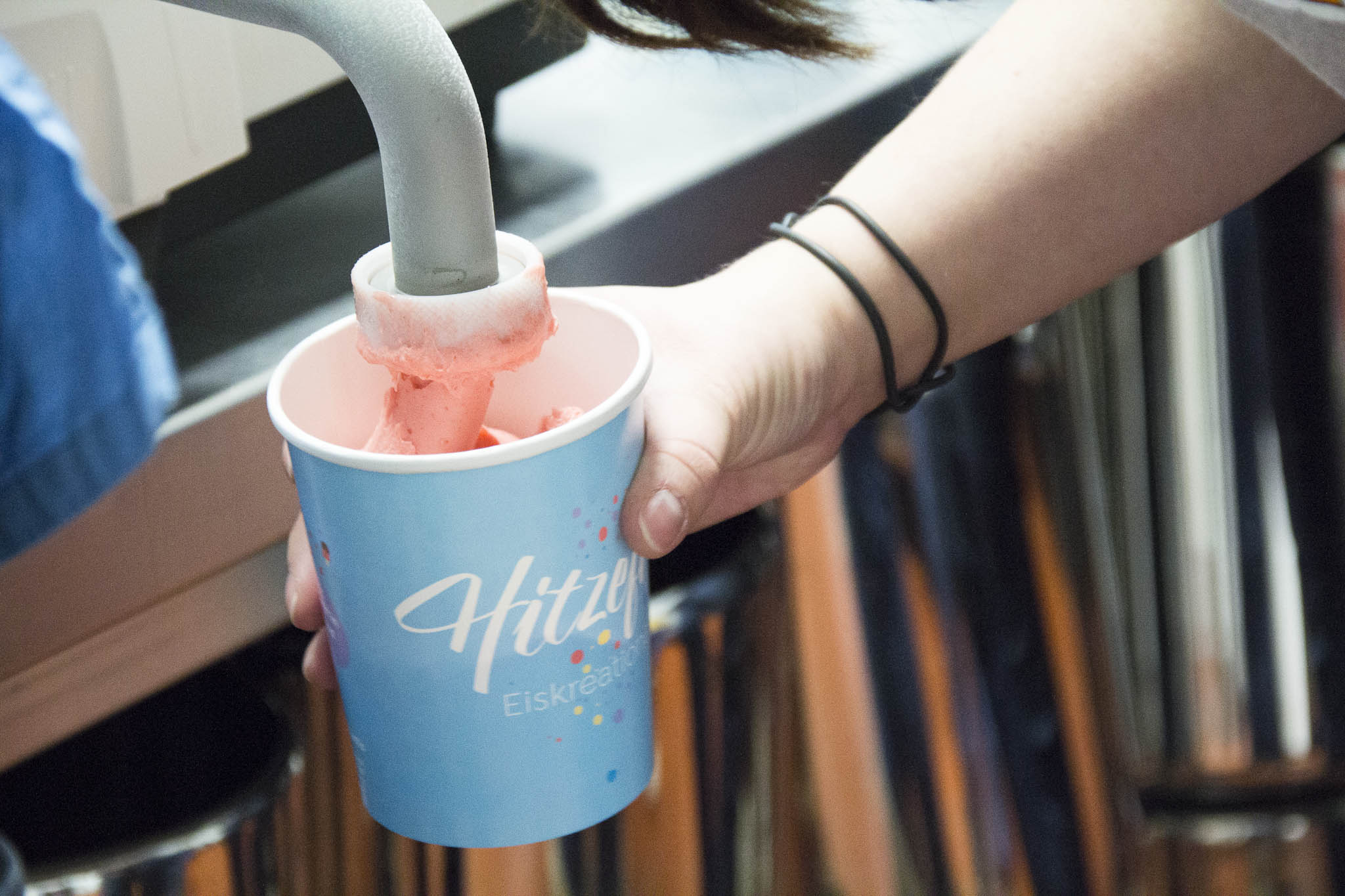 Eine Person hält einen hellblauen Becher mit rosa Speiseeis und Waffelstiel in der Hand.