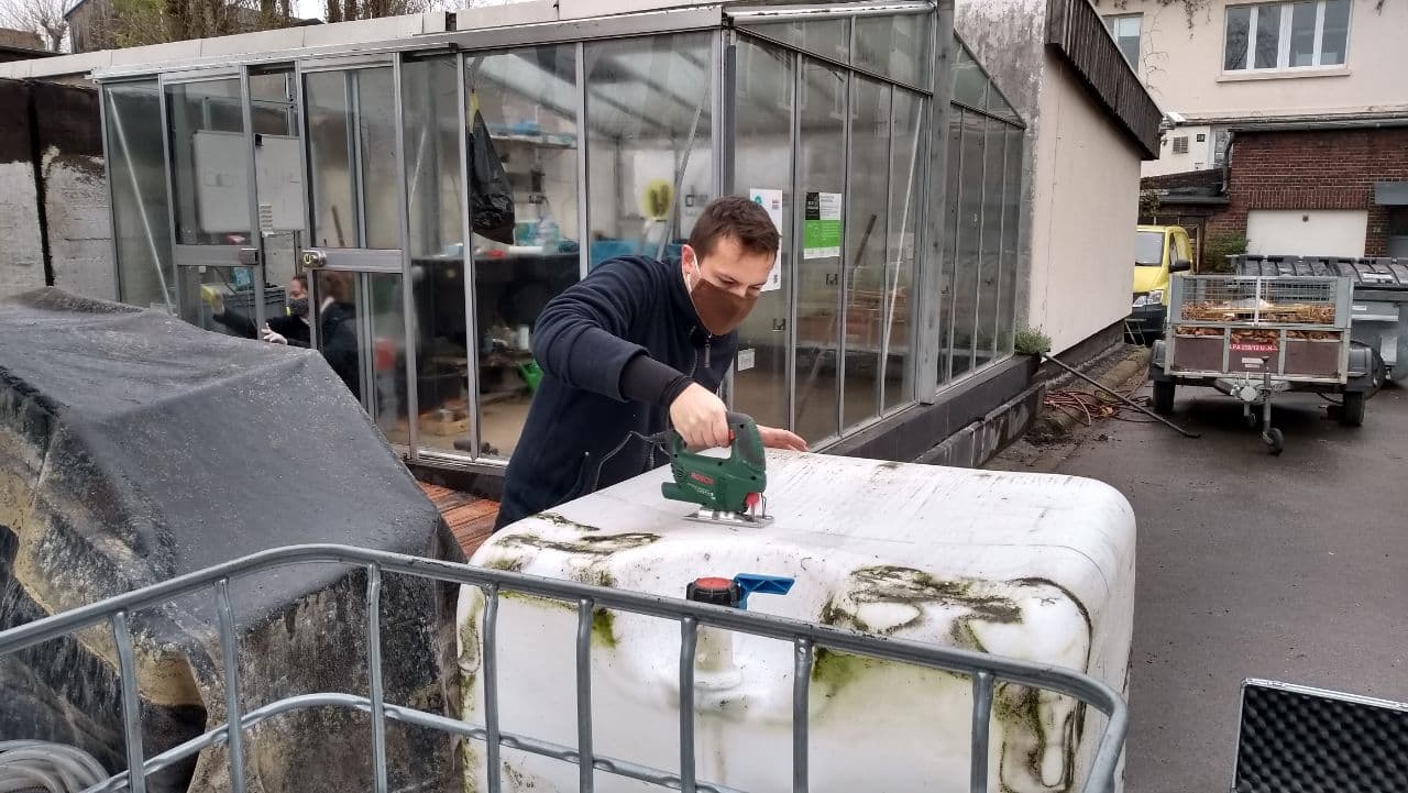 Ein Mann bearbeitet mit einer Säge ein weißes Objekt in einem Innenhofbereich neben Werkzeugen.