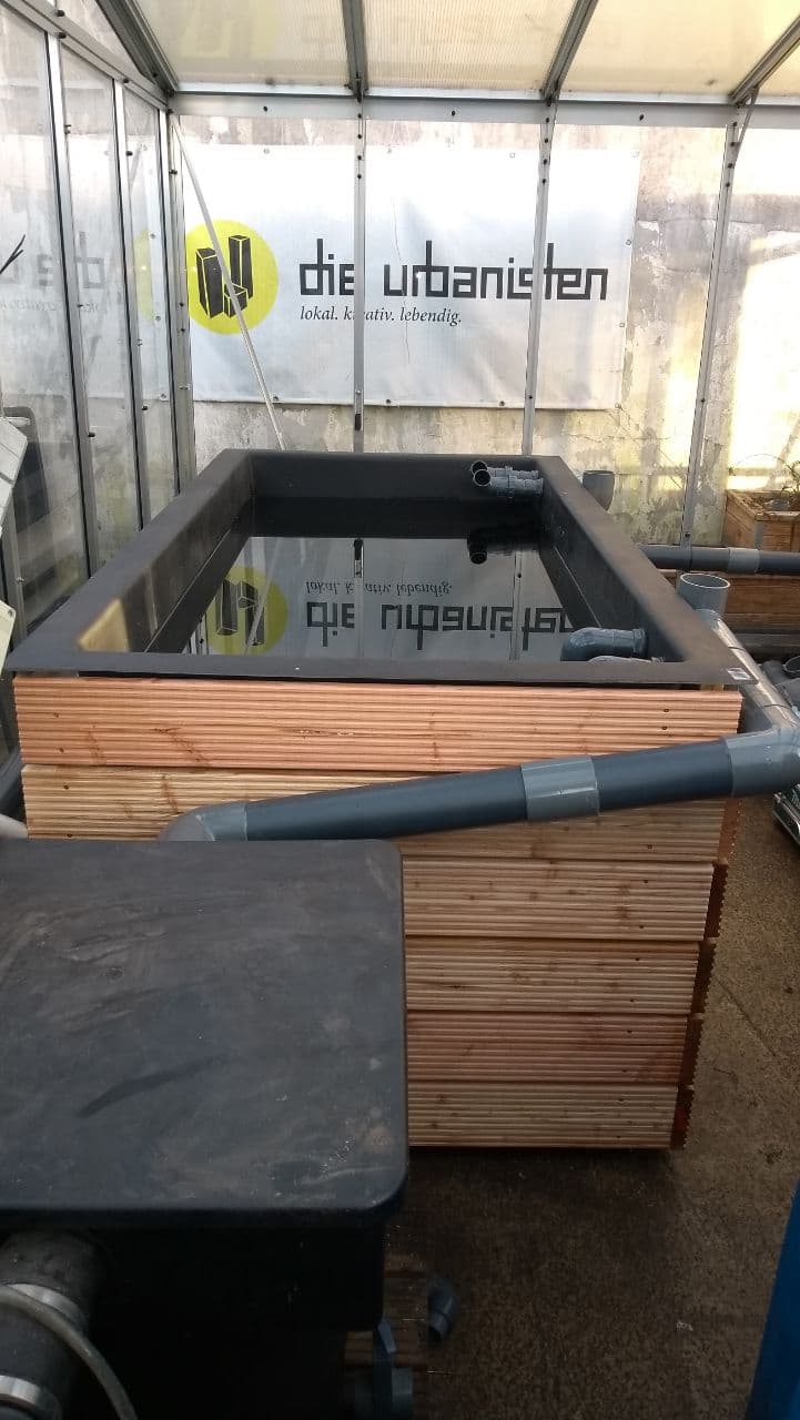 Hochbeet aus Holz und schwarzem Kunststoff in transparentem Gewächshaus mit Vereinslogo.