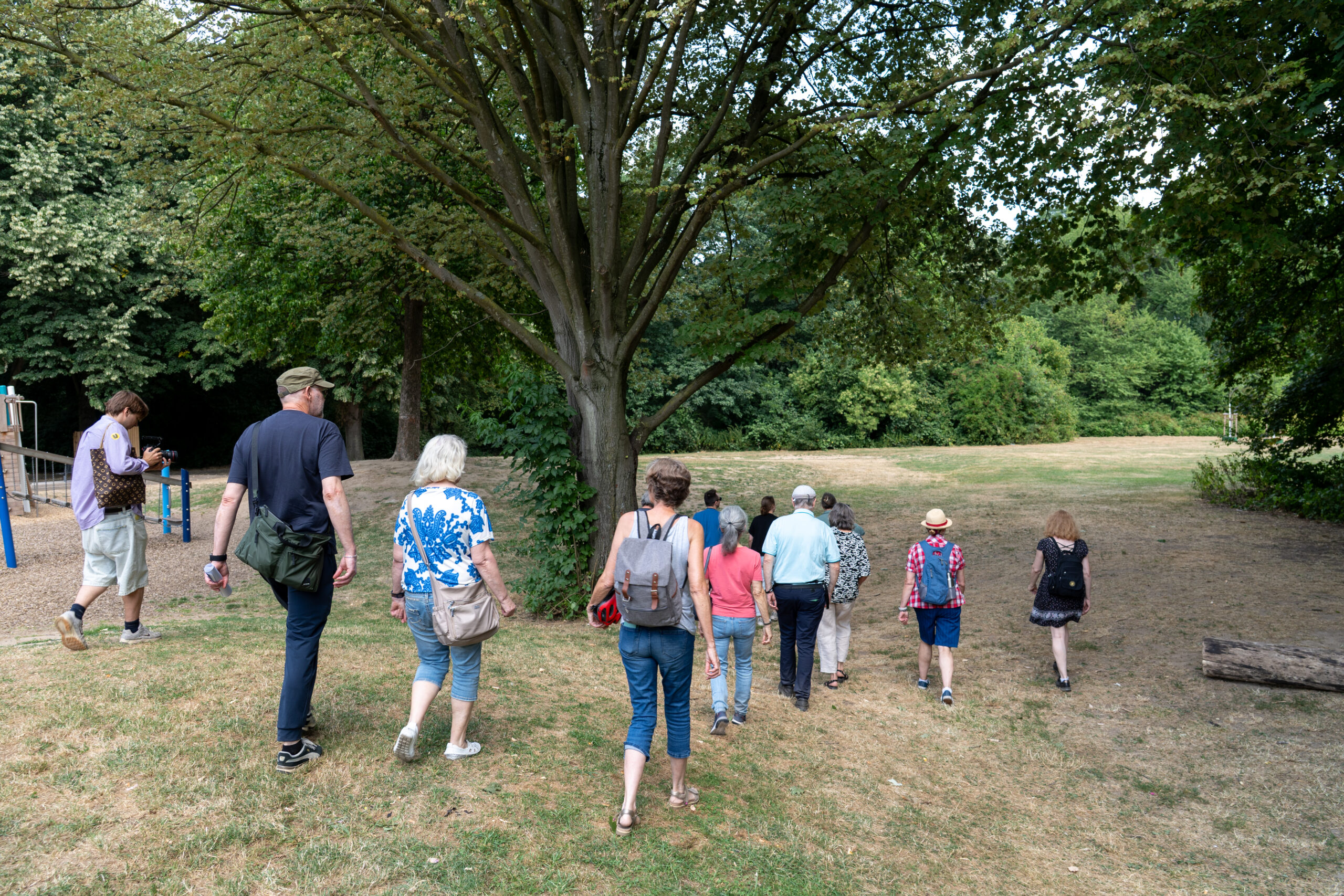 Eine Gruppe von Menschen verschiedenen Alters joggt gemeinsam durch einen grünen Park unter großen Bäumen.