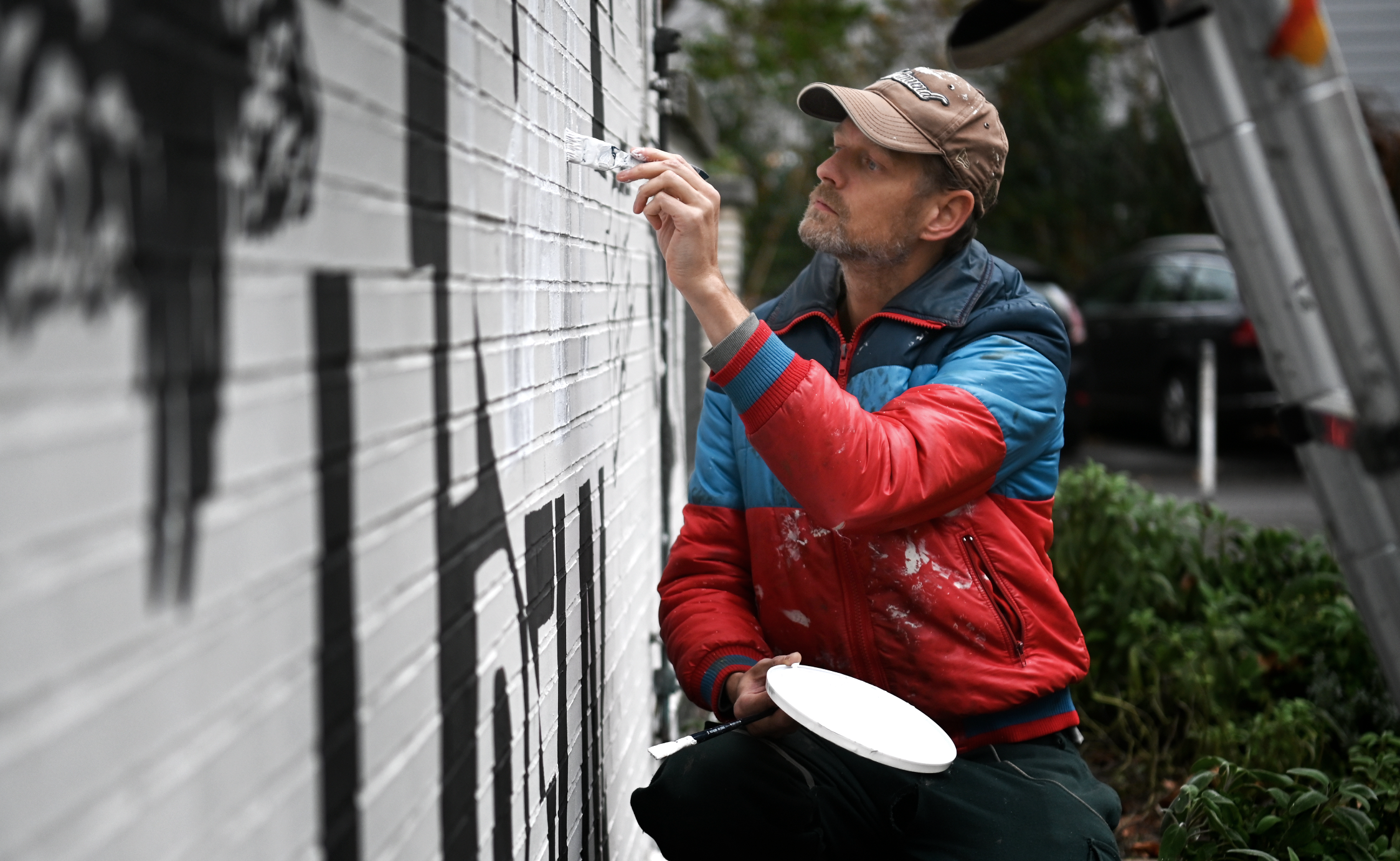Mann in roter Jacke bemalt weiße Wand mit Pinsel und Farbpalette.