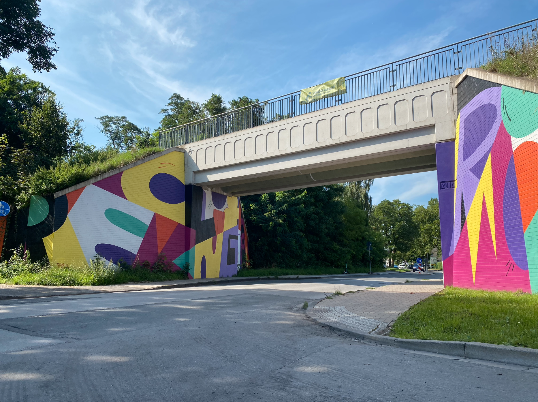 Betonbrücke mit bunten geometrischen Wandmalereien in Gelb, Pink, Lila und Orange an Brückenpfeilern.