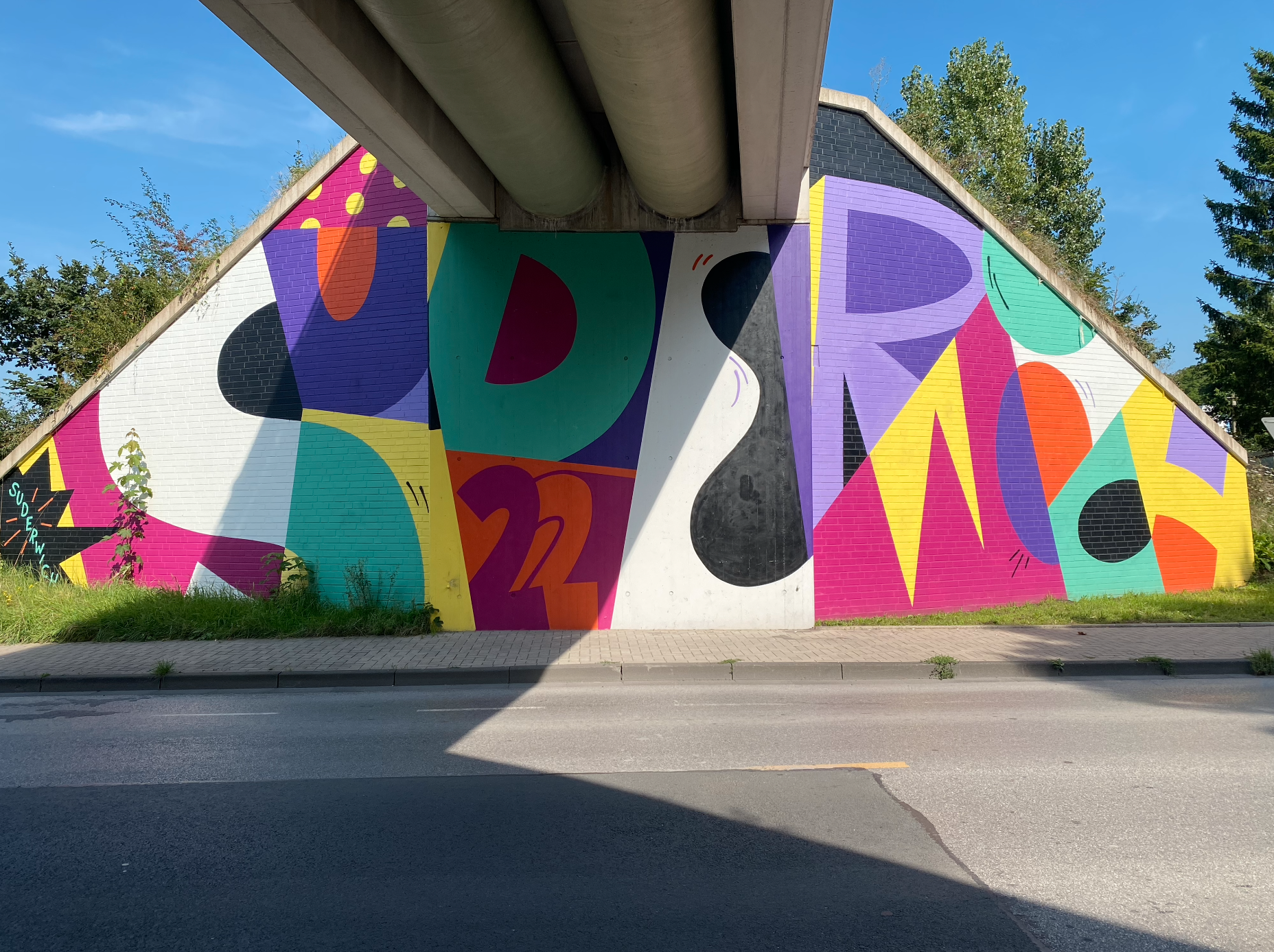 Buntes Wandgemälde mit geometrischen Formen unter einer Brücke an einer Straße.