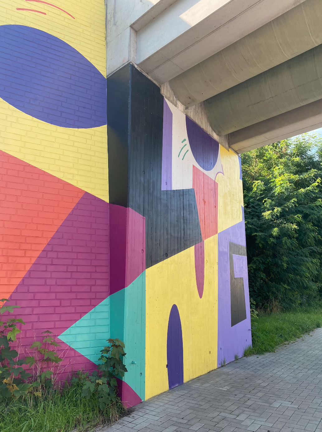 Großflächiges Wandgemälde unter Brücke mit geometrischen Formen in Gelb, Rot, Blau und Violett.