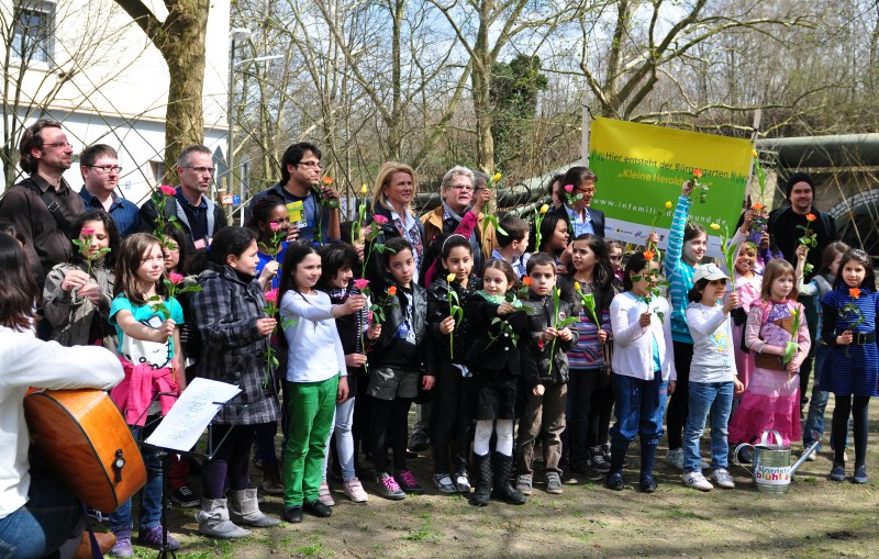 Eine Gruppe von Erwachsenen und Kindern steht outdoor mit Blumen und Transparenten zusammen.