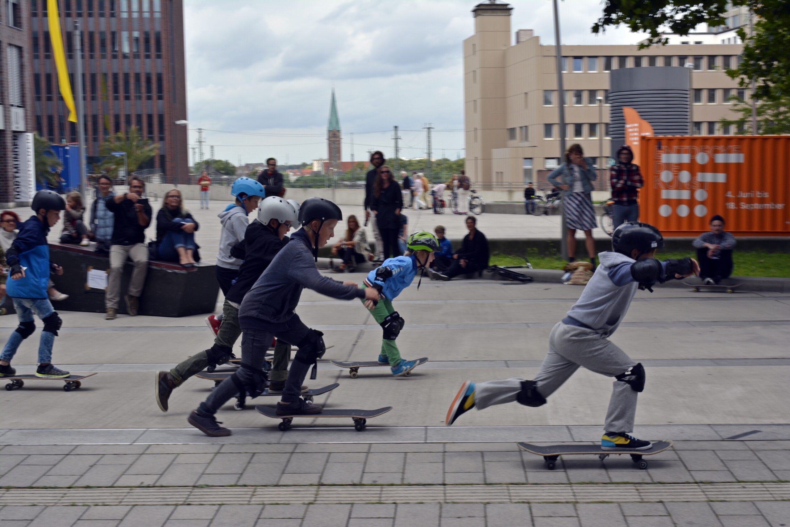 Mehrere Skateboardfahrer verschiedenen Alters trainieren auf einer befestigten Platzfläche mit Publikum.