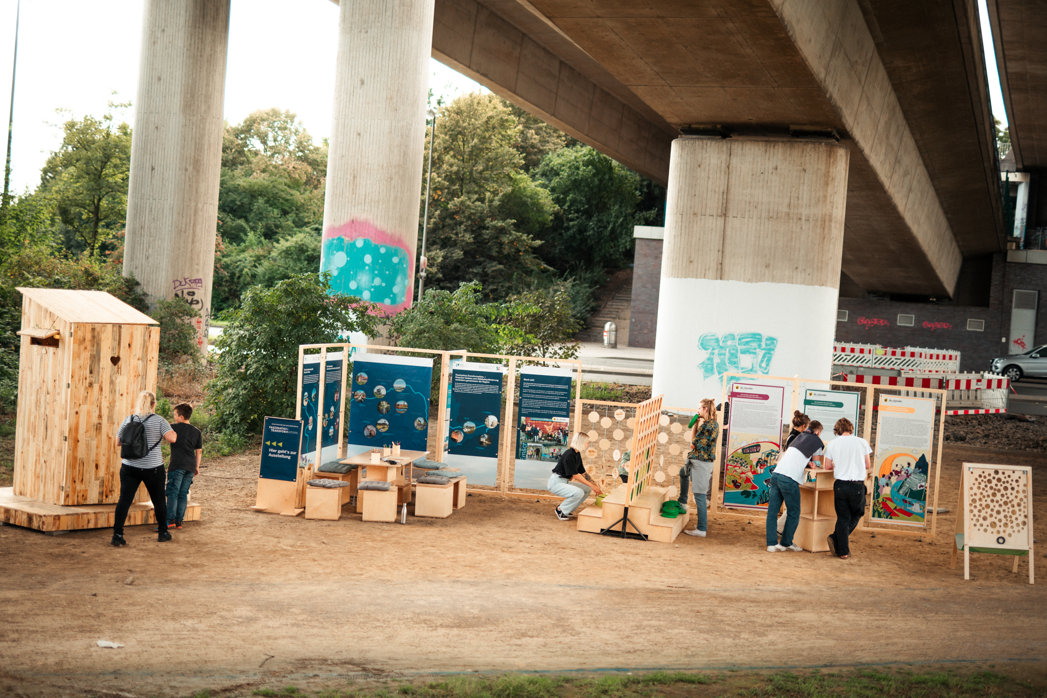 Unter einer Autobahnbrücke präsentieren mehrere Personen Ausstellungstafeln und Holzstrukturen an einem Sandplatz.