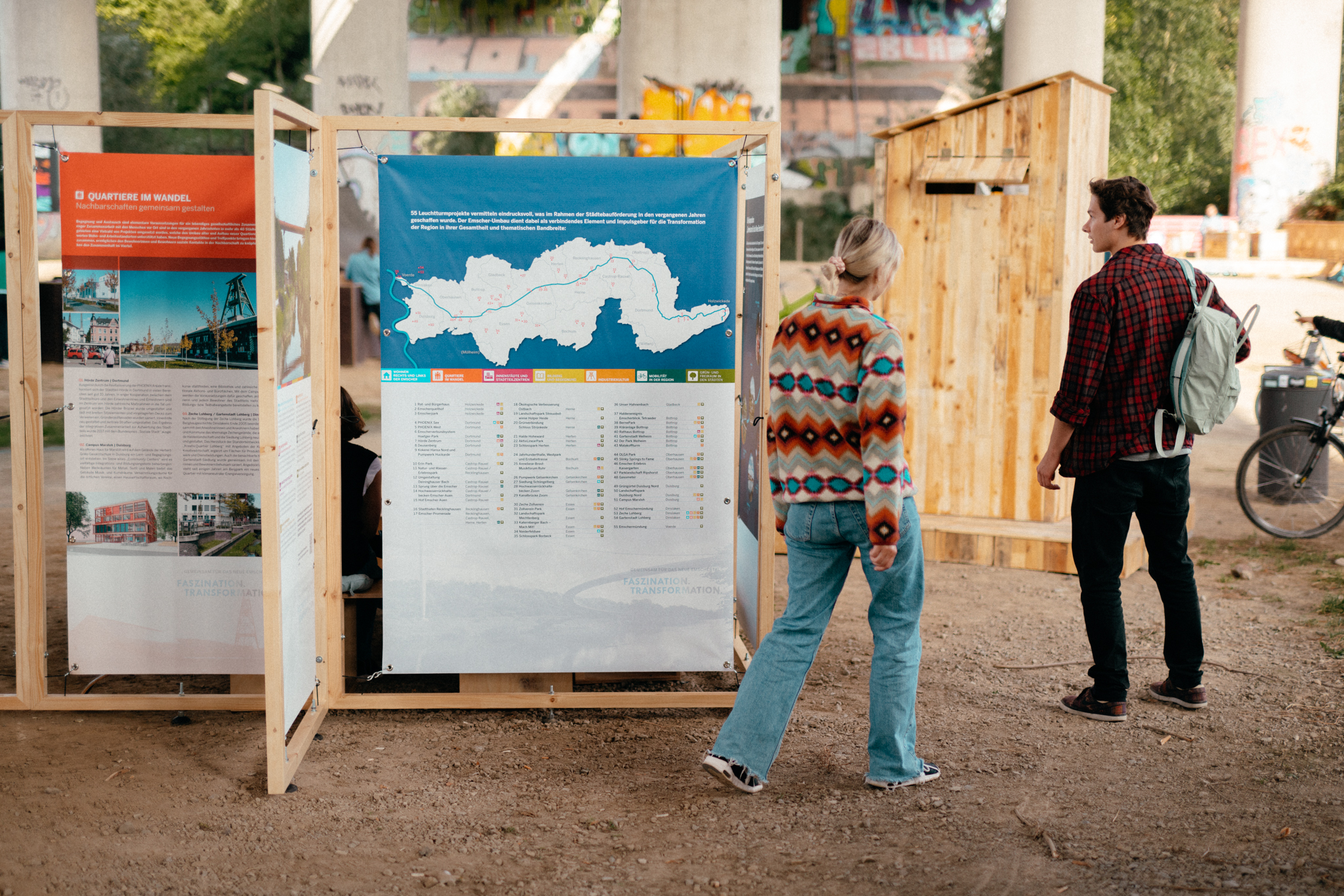 Zwei Personen betrachten Informationstafeln zu Stadtentwicklung an einer Ausstellung unter einer Brücke.