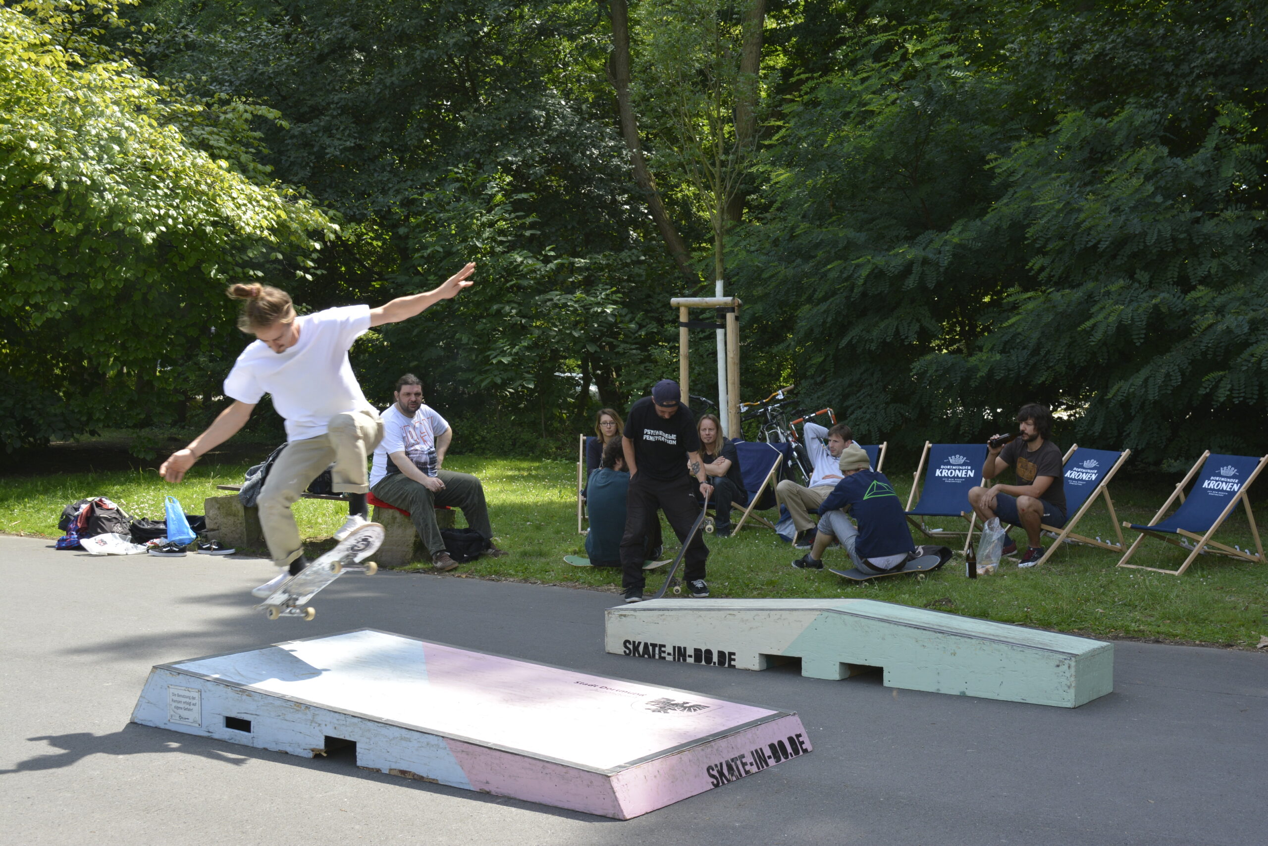 Skateboarder führt Trick auf Rampe vor sitzendem Publikum in Parkanlage auf.