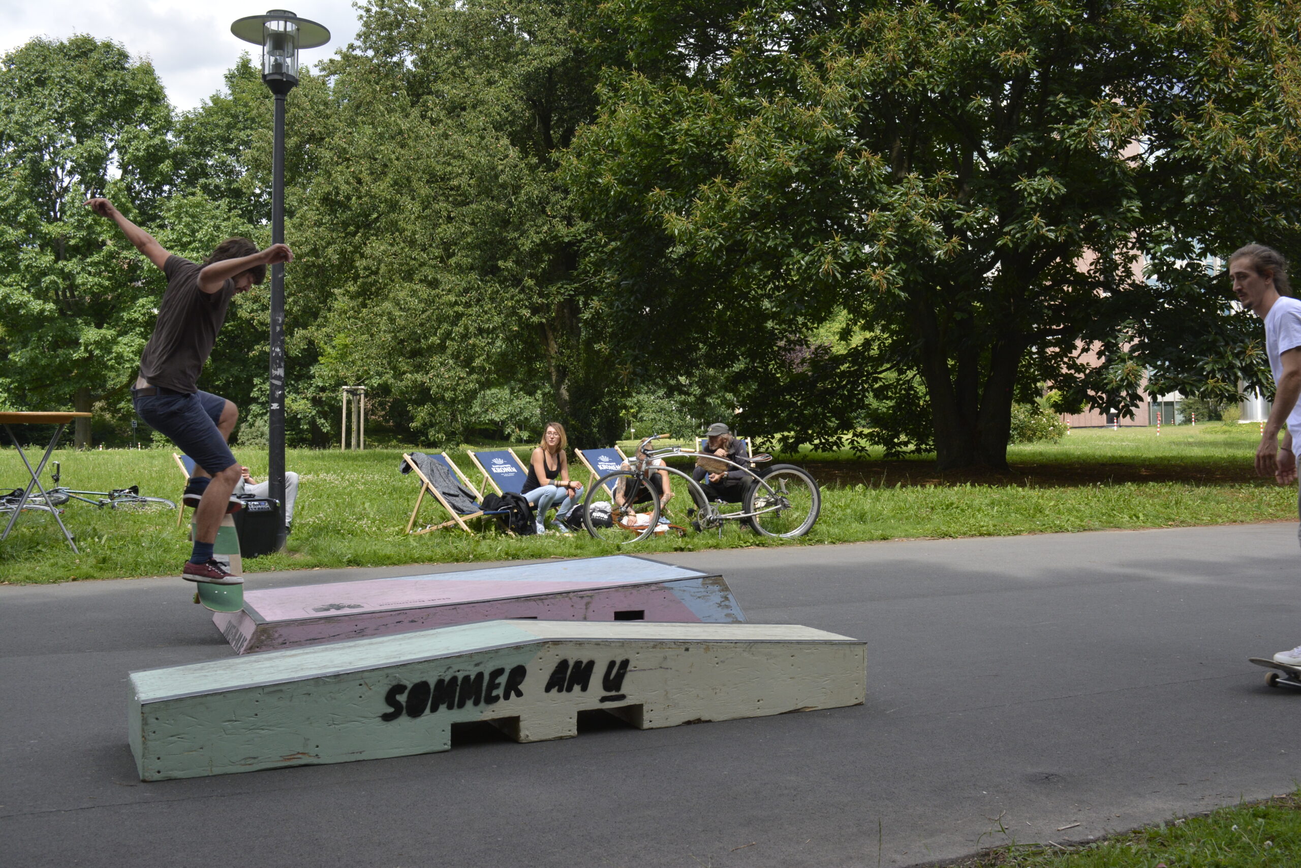 Ein Skateboarder springt über ein bemaltes Hindernis in einem Park mit Zuschauern.