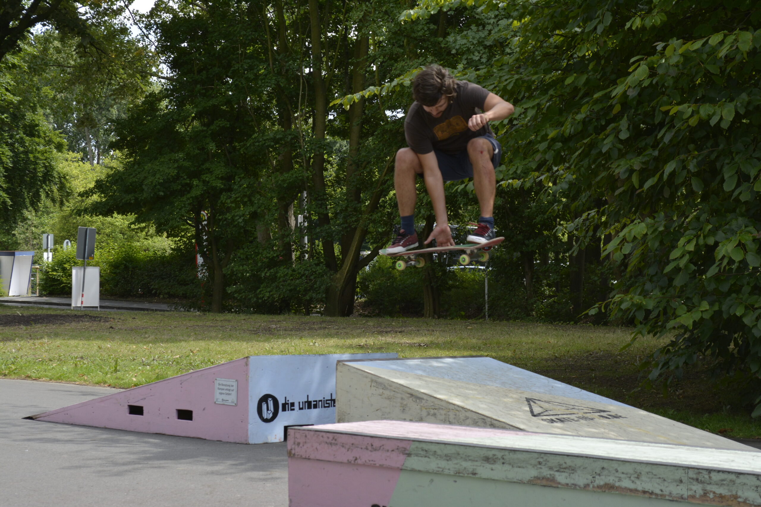 Ein Skateboarder springt über eine Rampe in einem Park mit Bäumen.