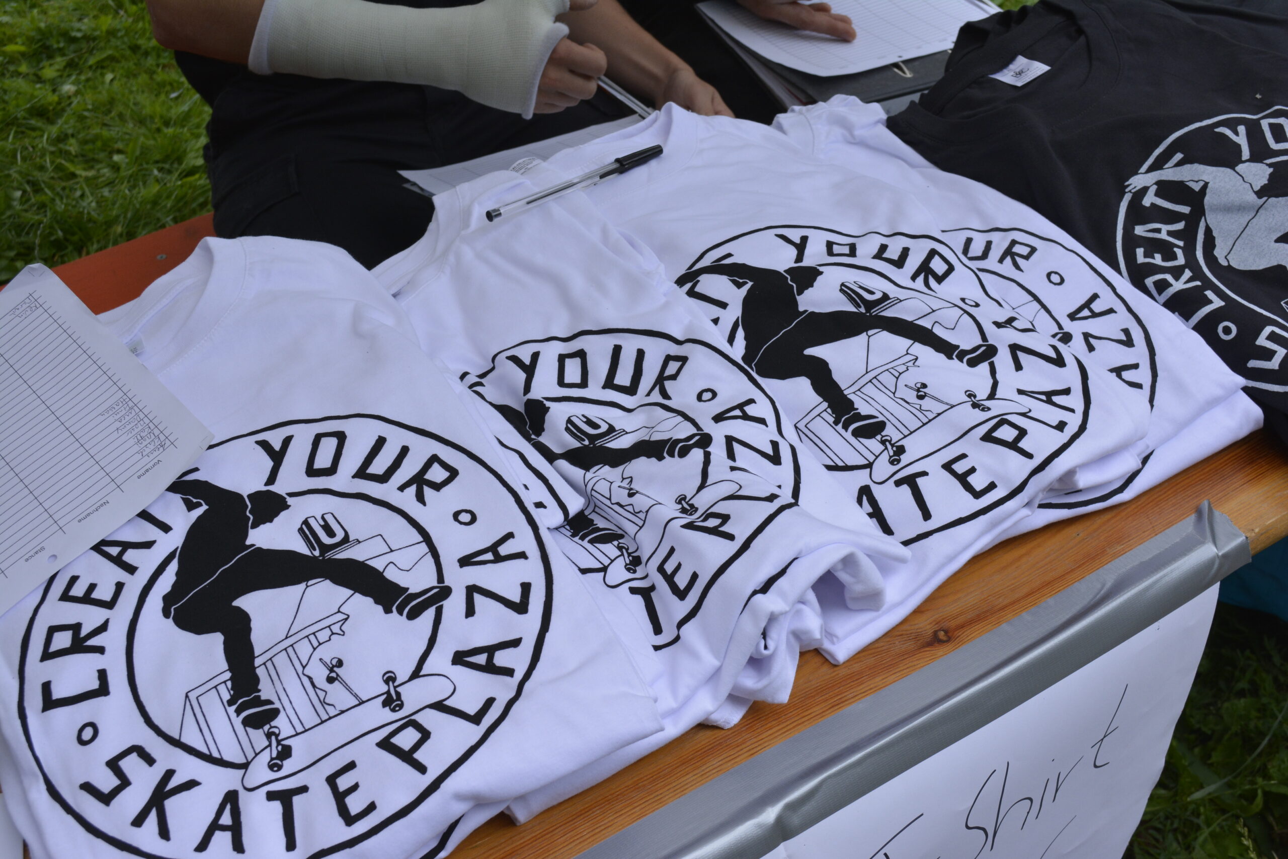Drei weiße T-Shirts mit Skatepark-Design liegen auf Holztisch neben Dokumenten.