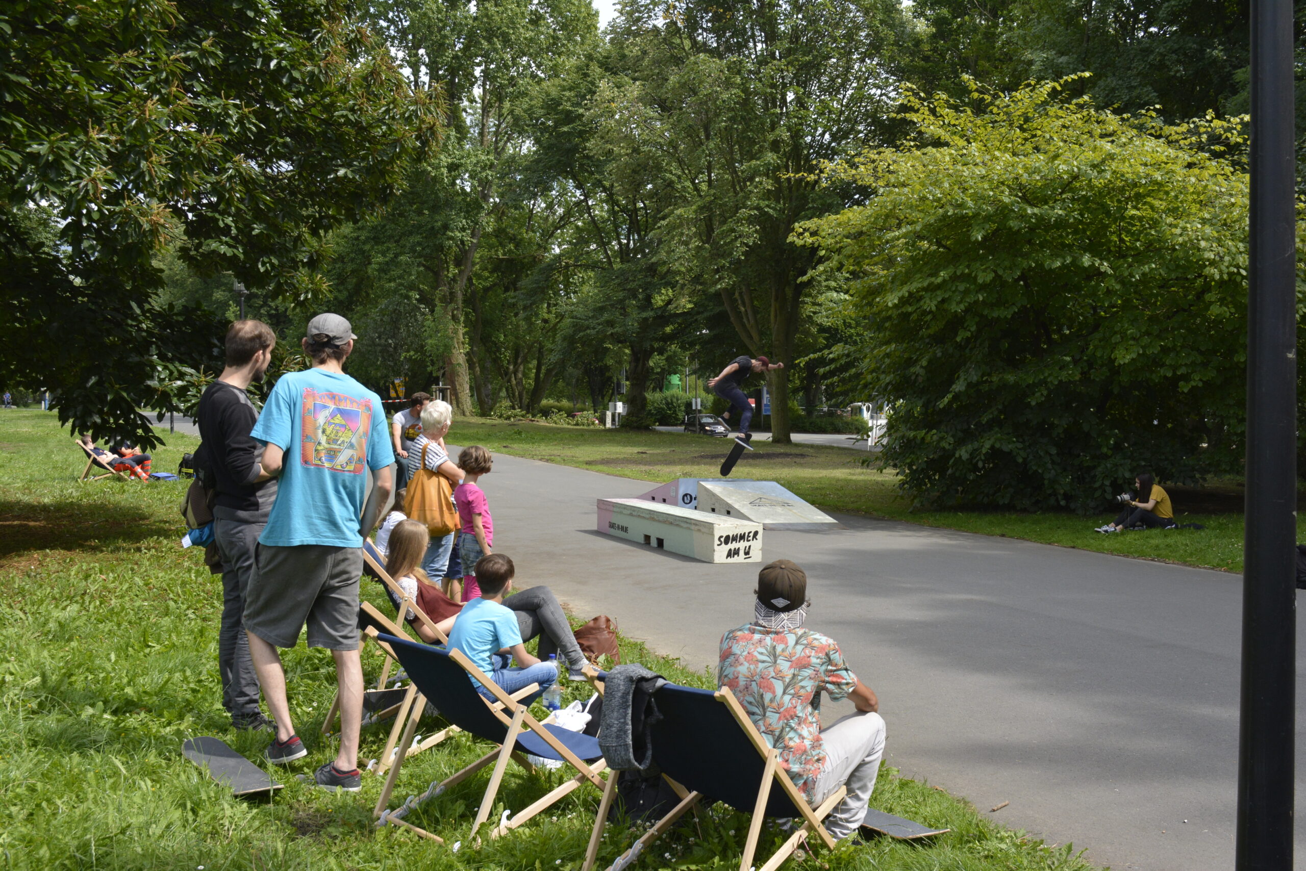 Familie sitzt auf Stühlen am Rand eines Skate-Parks und beobachtet Skateboard fahrende Personen.