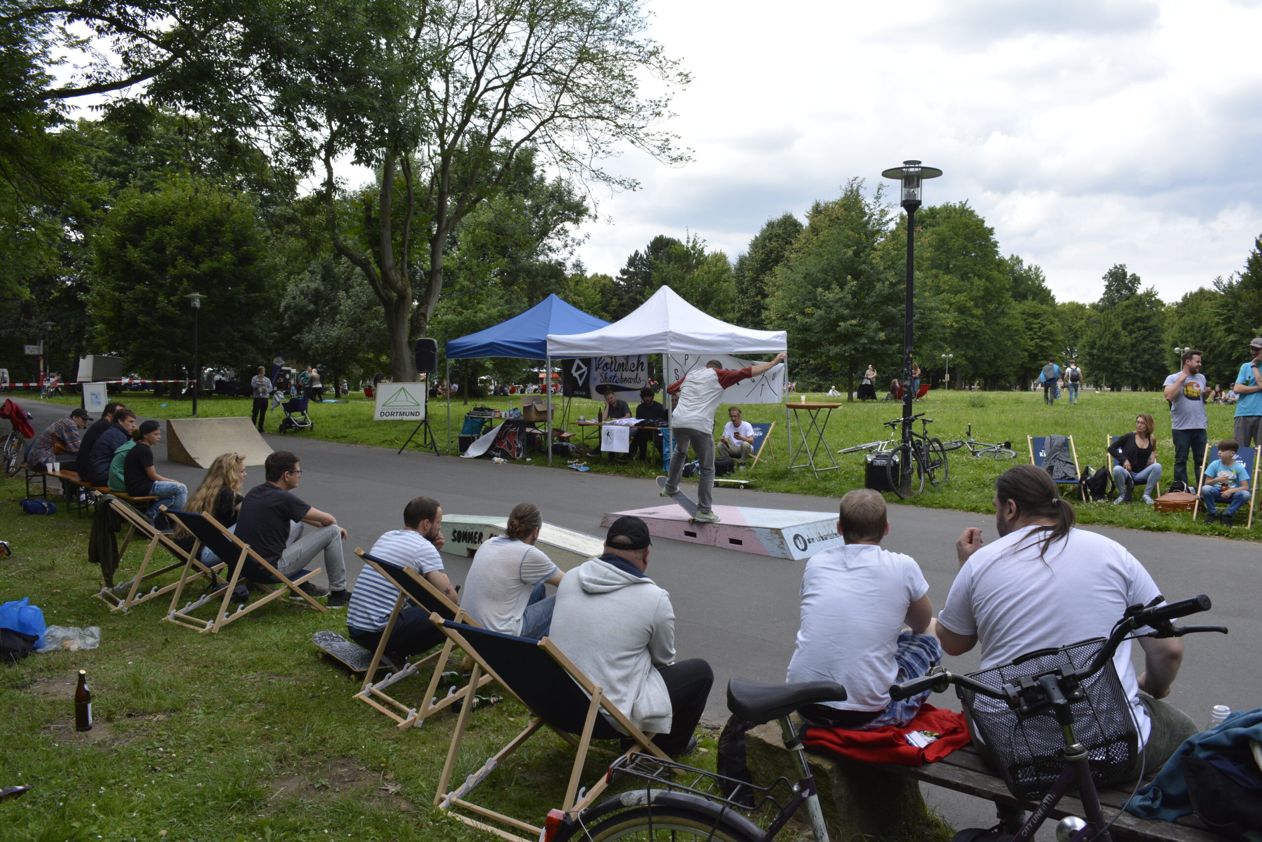 Sportplatz in Park mit Skateboard-Rampe, Zuschauern auf Liegestühlen und Zeltpavillons.