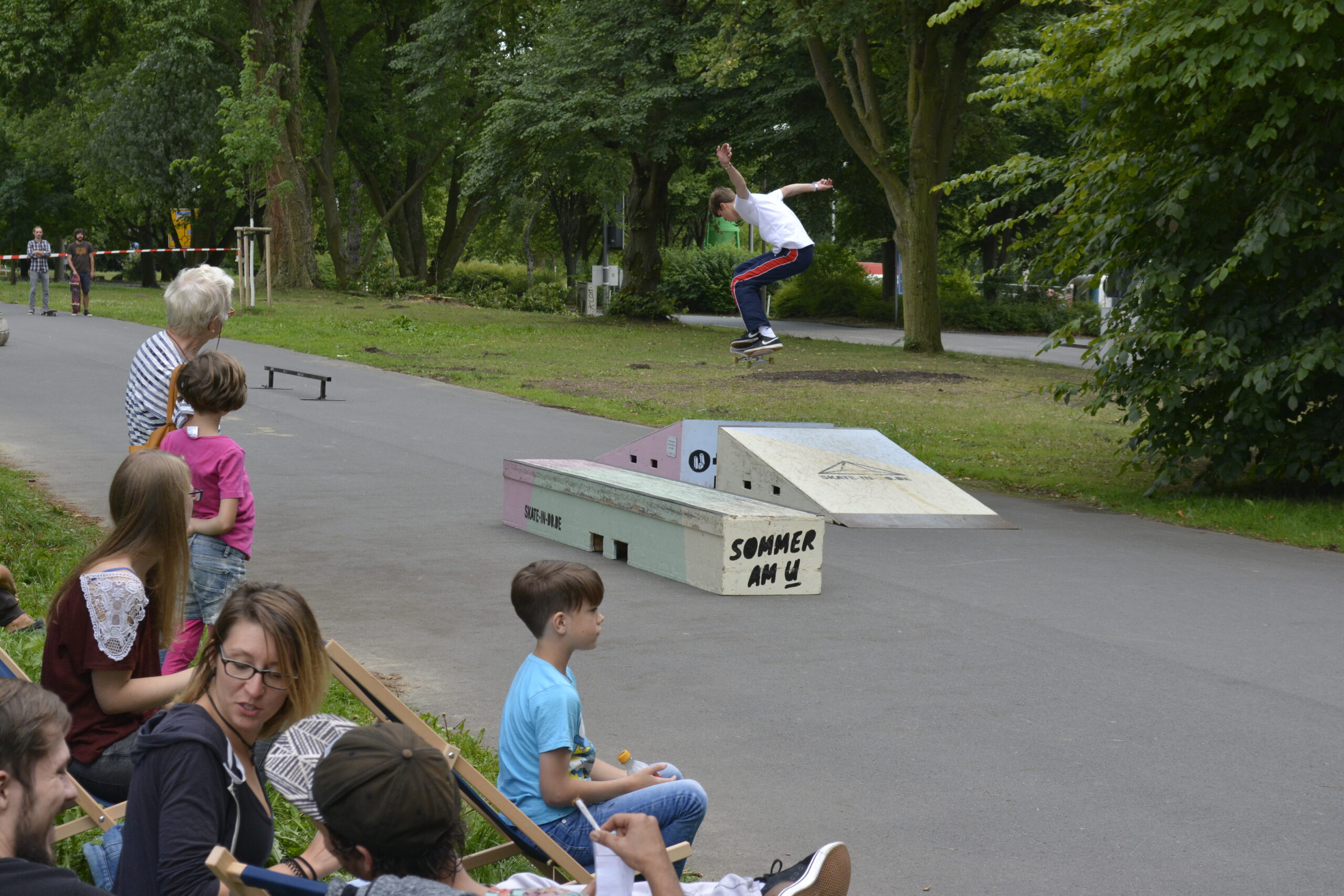 Skateboardfahrer springt über Hindernis in Skatepark, Zuschauer beobachten von Bänken.