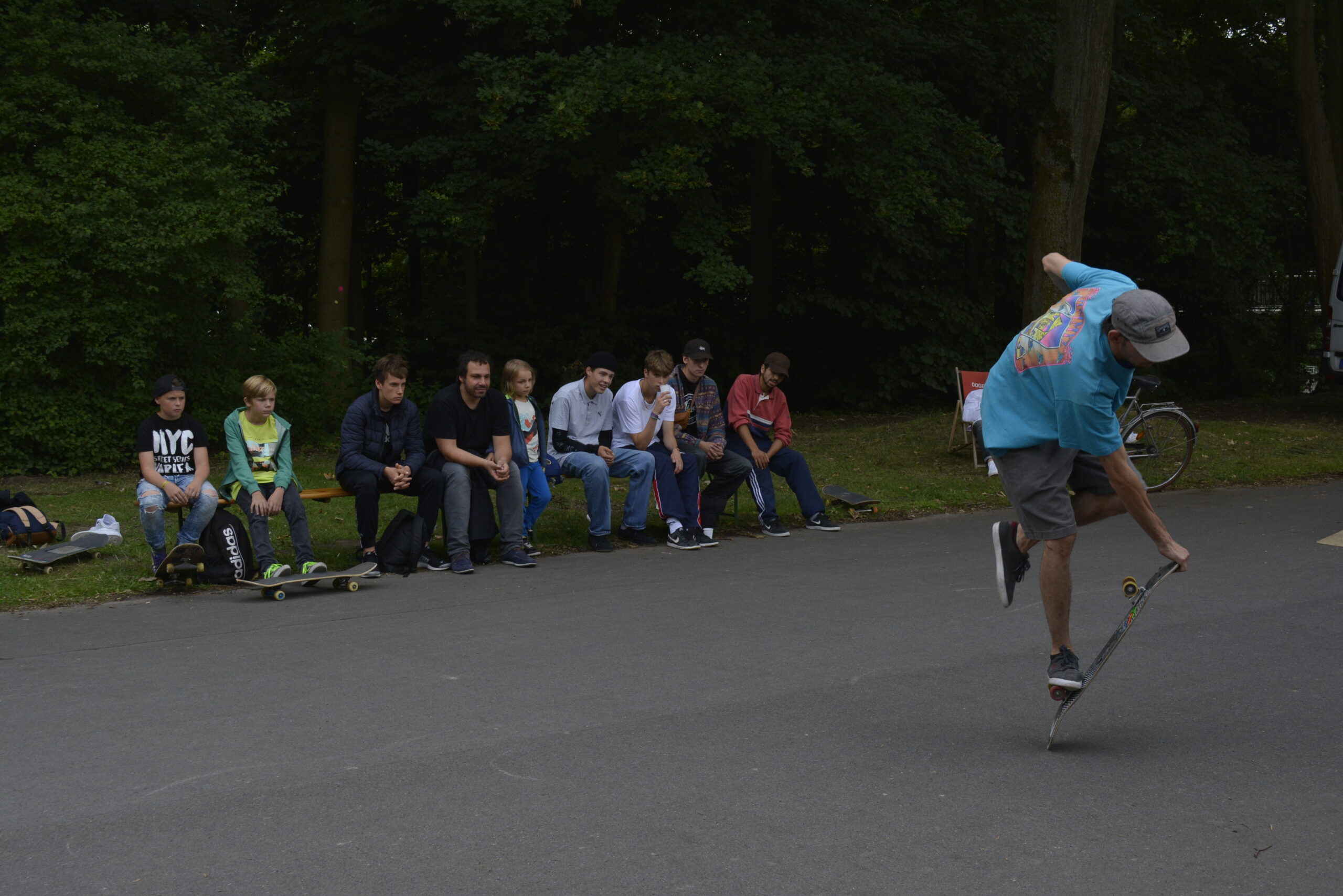 Ein Skateboarder führt Tricks vor, während eine Gruppe Jugendlicher auf einer Bank zusieht.