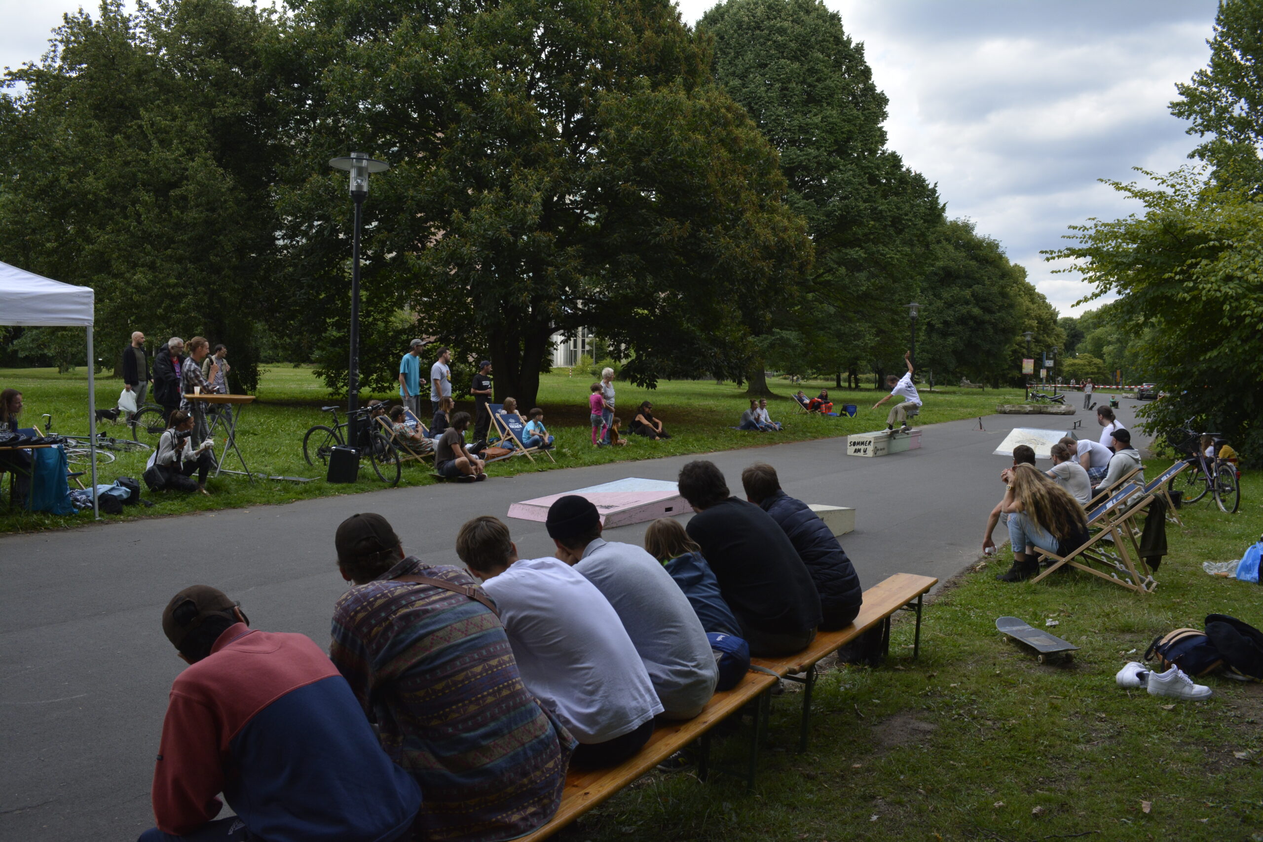 Mehrere Menschen sitzen auf Bänken und beobachten Skatboardfahrer auf einem Platz in einem Park.