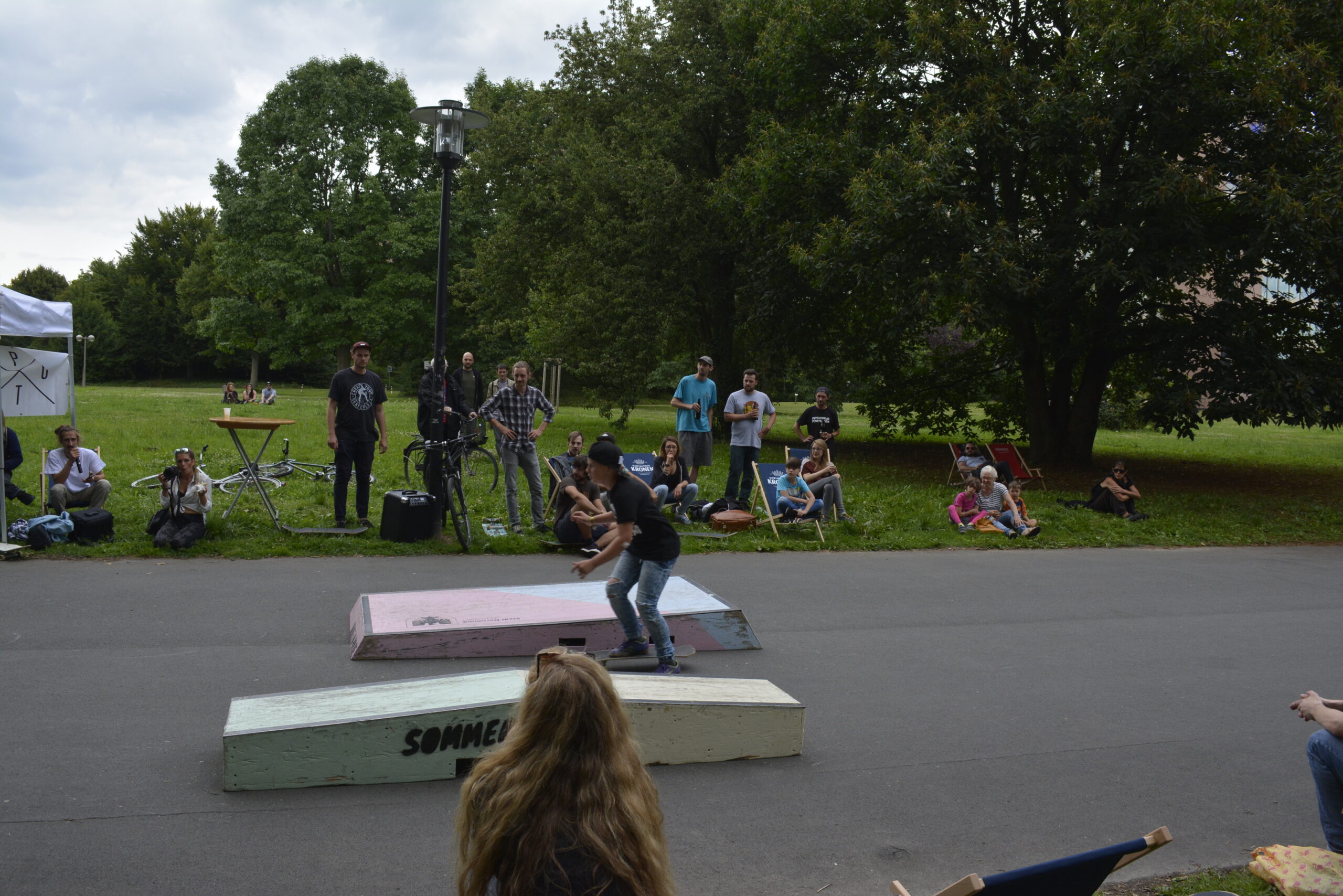 Skaterin springt über Rampe in Skatepark, Zuschauer sitzen auf Rasen im Hintergrund.