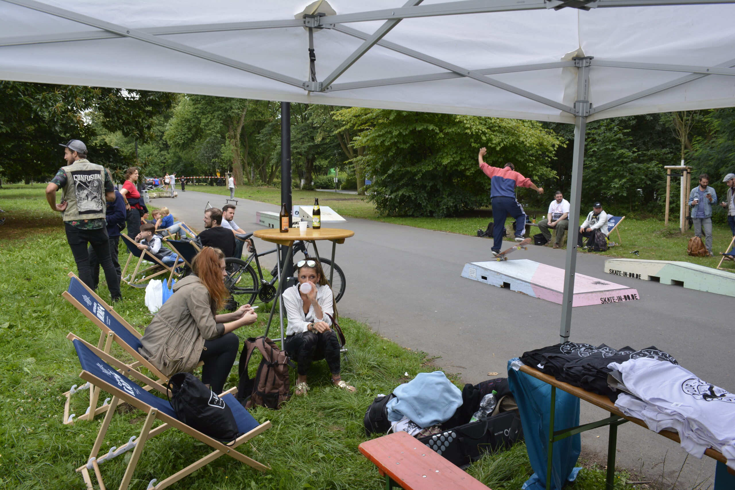 Skateboard-Event in einem Park mit Zuschauern unter Zelt und Fahrer auf der Rampe.