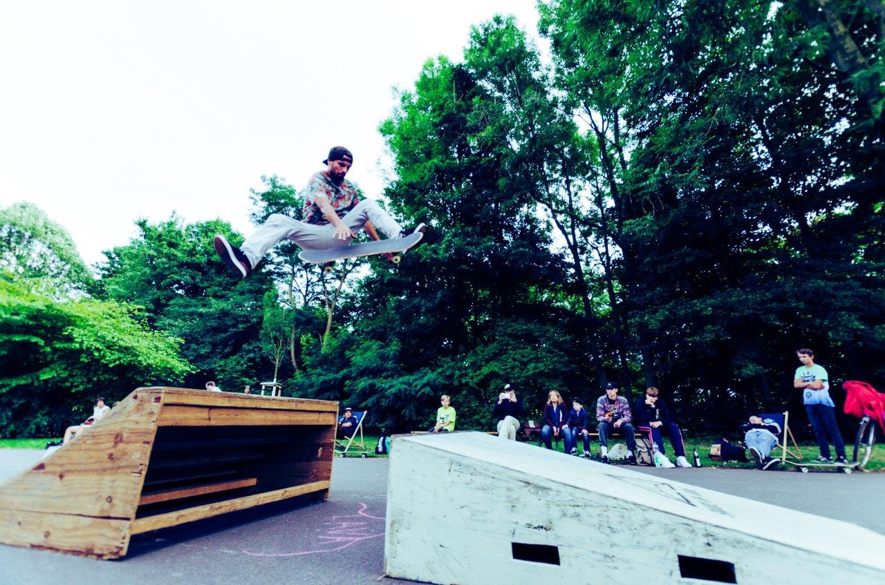 Ein Mann springt über Hindernisse auf einem Skatepark vor Zuschauern.