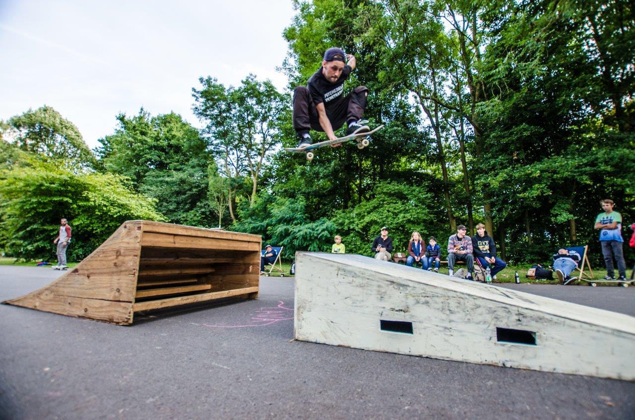 Ein Skateboarder springt über ein weißes Betonobstacle in einem Skatepark, Zuschauer sitzen daneben.
