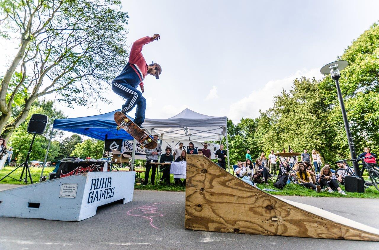 Skateboarder springt über Holzrampe bei Ruhr Games Event im Park vor Publikum.