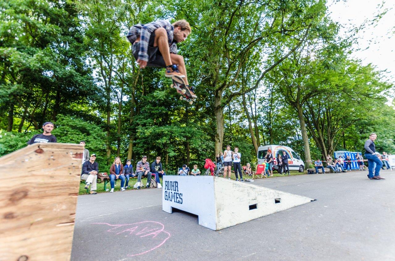 Ein Skateboarder springt über ein Hindernis auf einer Rampe vor Zuschauern.