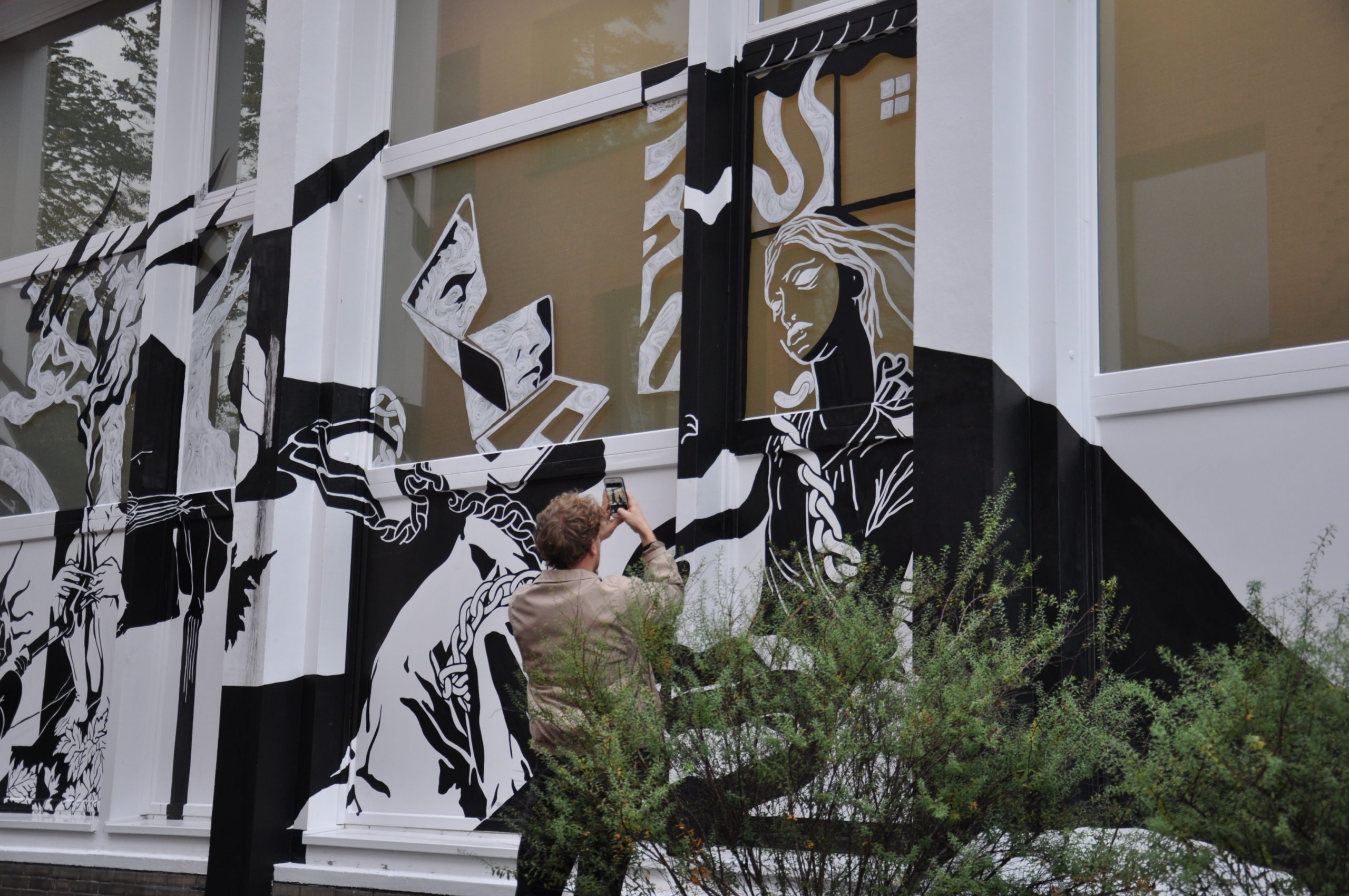 Person fotografiert schwarzweiße Wandmalerei mit Porträts an Hausfassade.