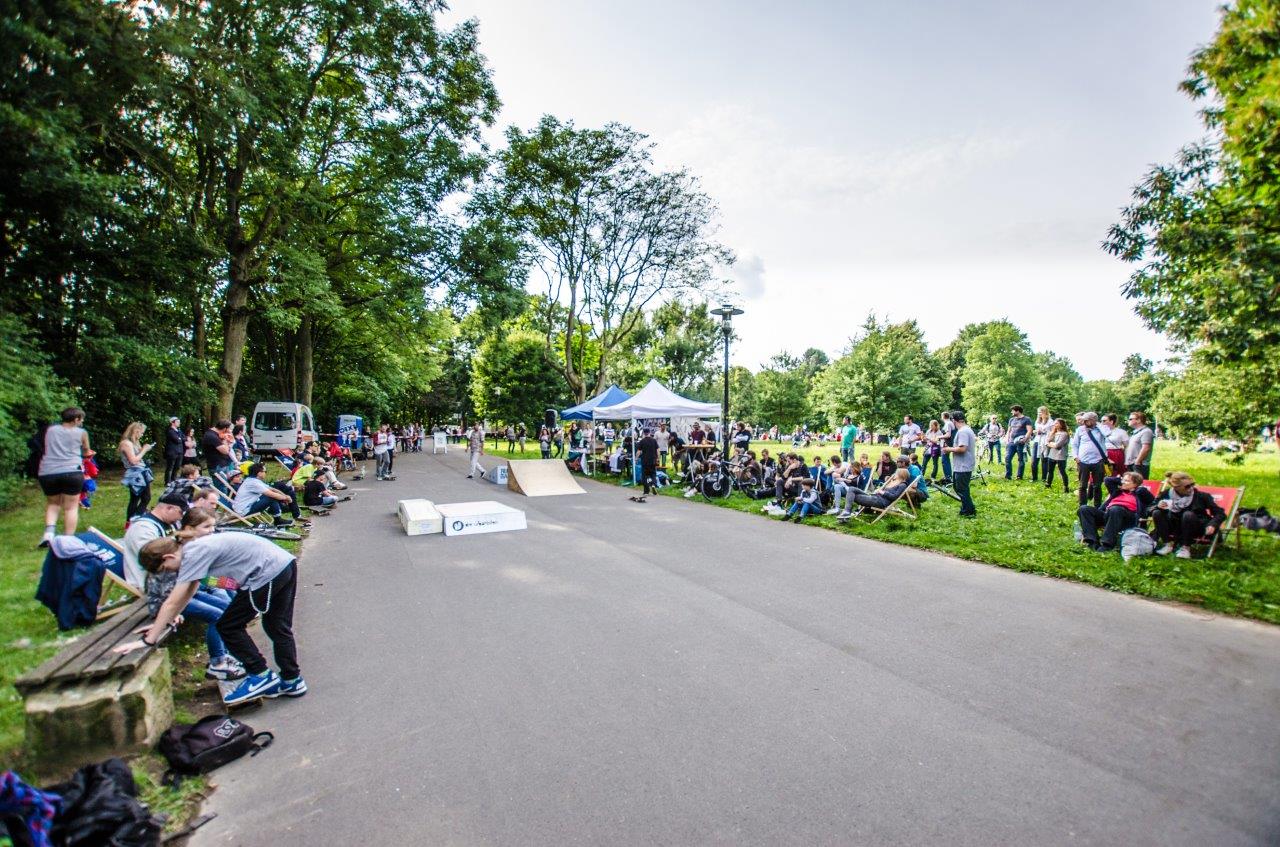 Öffentlicher Park mit Skateboard-Event: Zuschauer sitzen auf Rasen, Skateboarder nutzen Rampe, Zelt im Hintergrund.