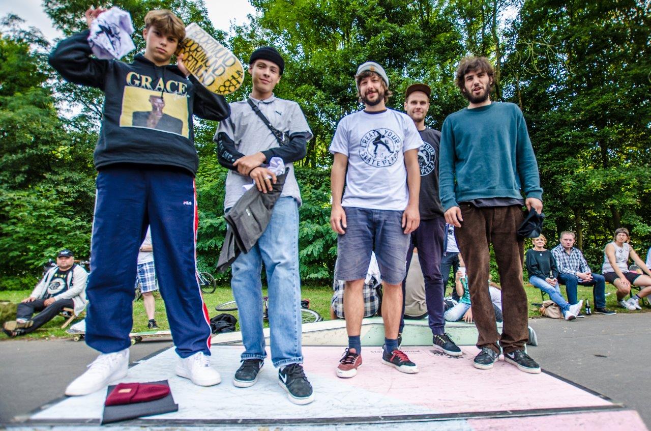 Fünf Personen stehen auf einer Skate-Rampe in einem Park vor Zuschauern.