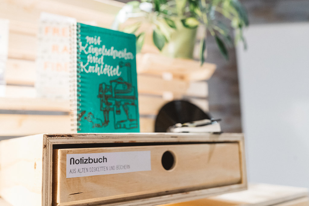 Holzboxen mit Notizbüchern und grünem Spiralblock auf Holztisch mit Pflanze.