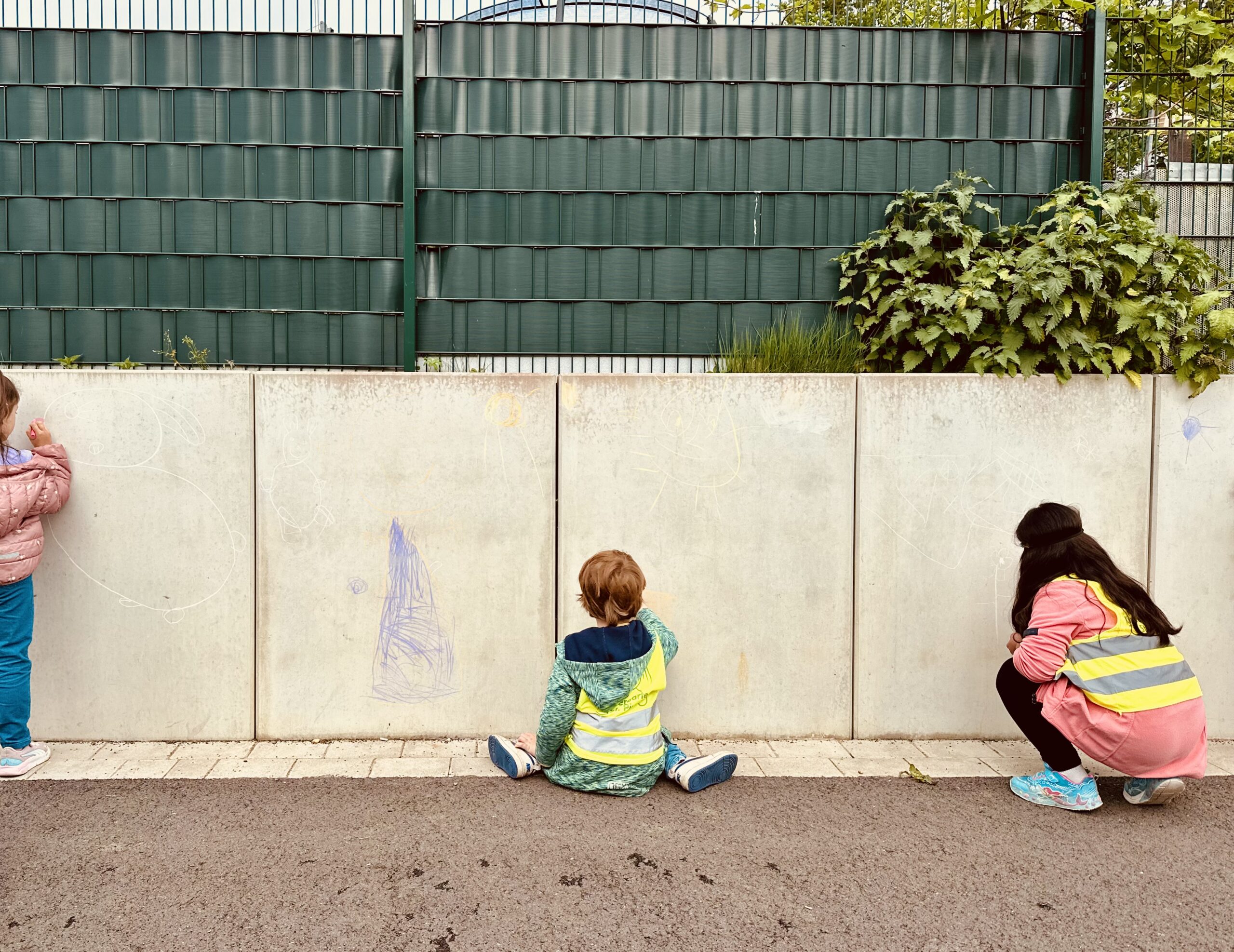Kinder zeichnen und malen an einer hellen Betonwand in einem Außenbereich.