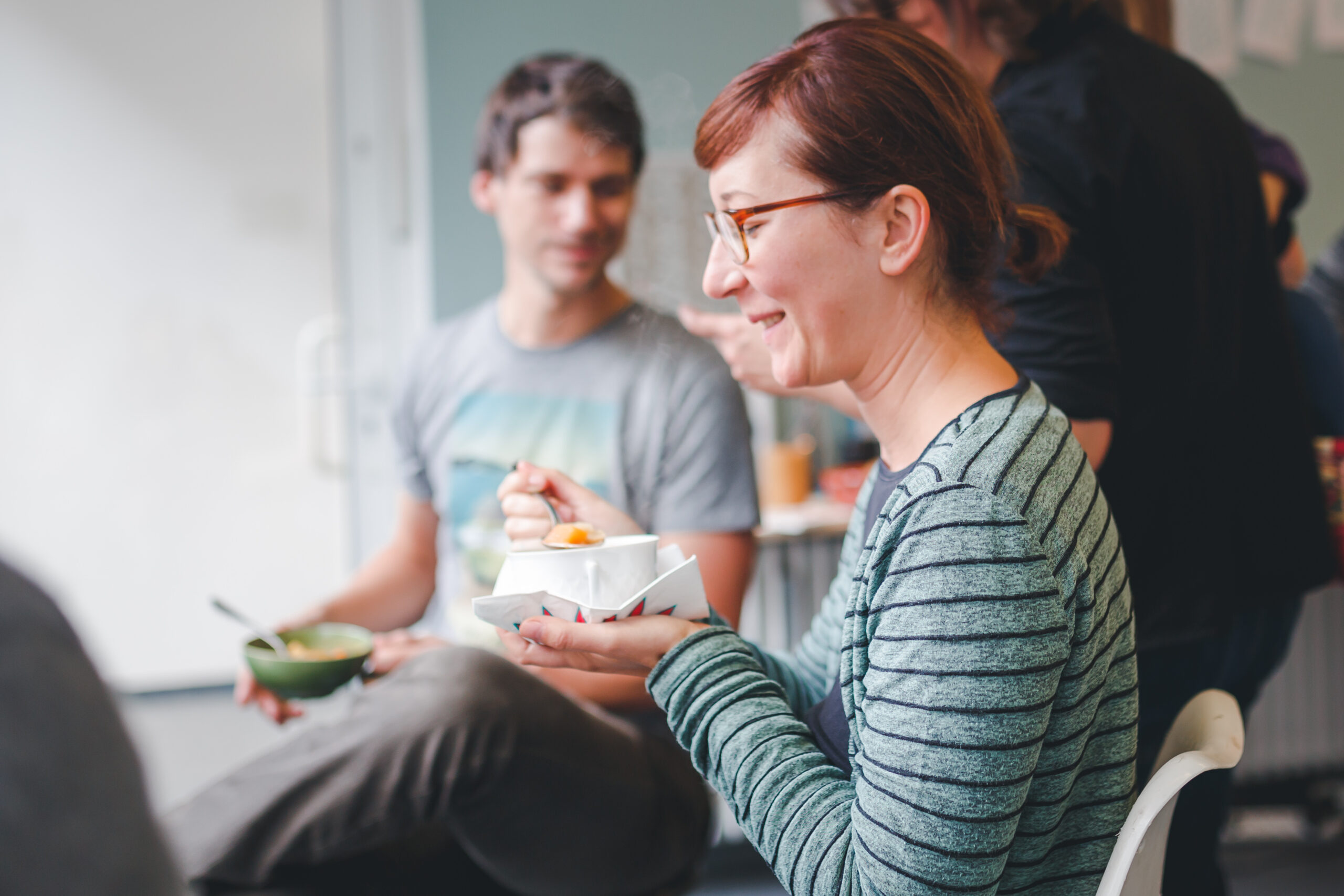 Eine Frau mit Brille hält einen Behälter und sitzt in einer Gruppe von Menschen bei einem gemeinsamen Essen.