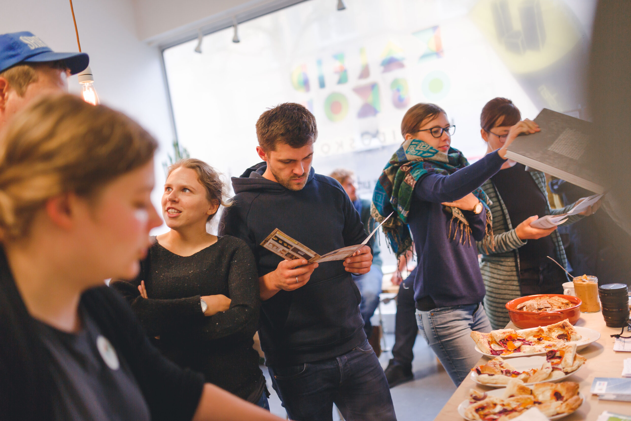 Gruppe von Menschen in Innenraum mit Pizza und Getränken beim gemeinsamen Austausch.