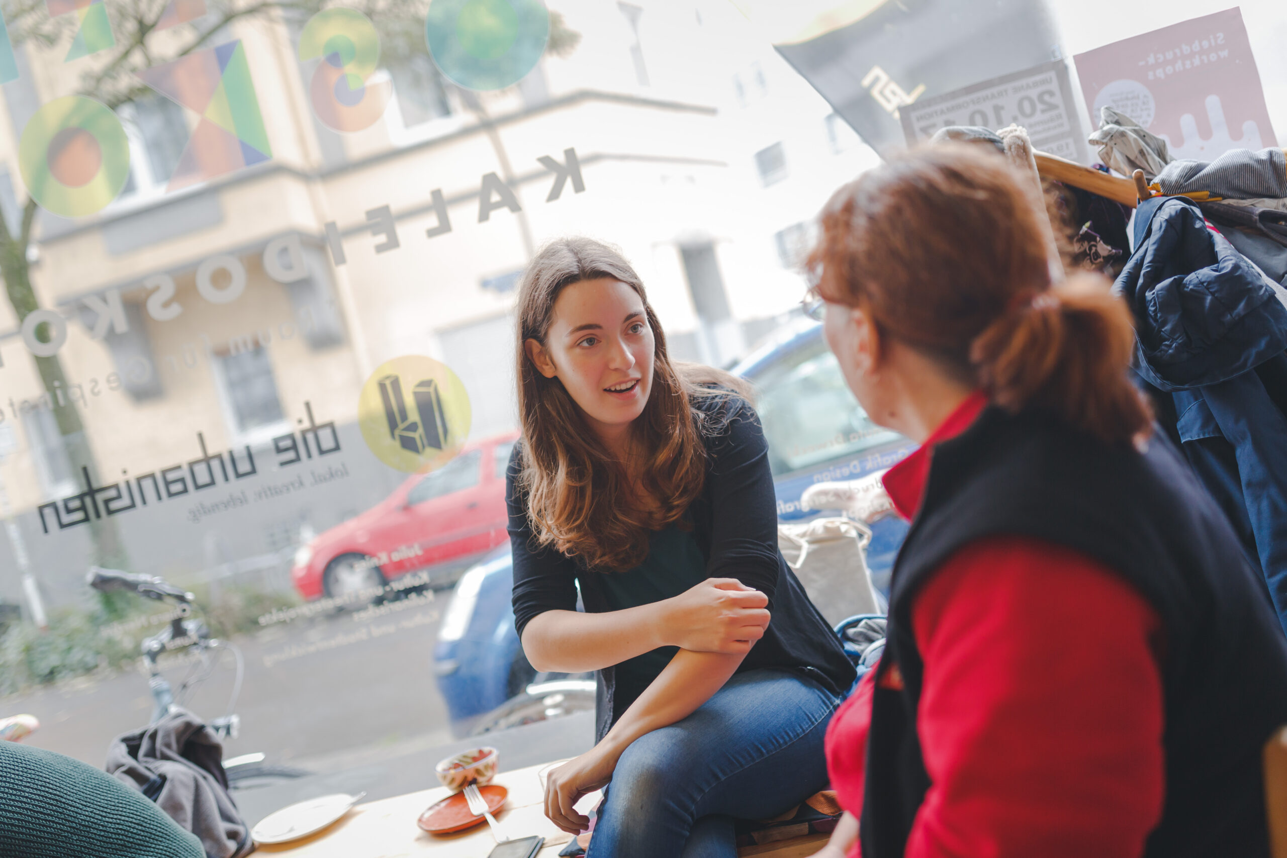 Zwei Frauen sitzen sich in einem urbanen Außenbereich gegenüber und unterhalten sich.