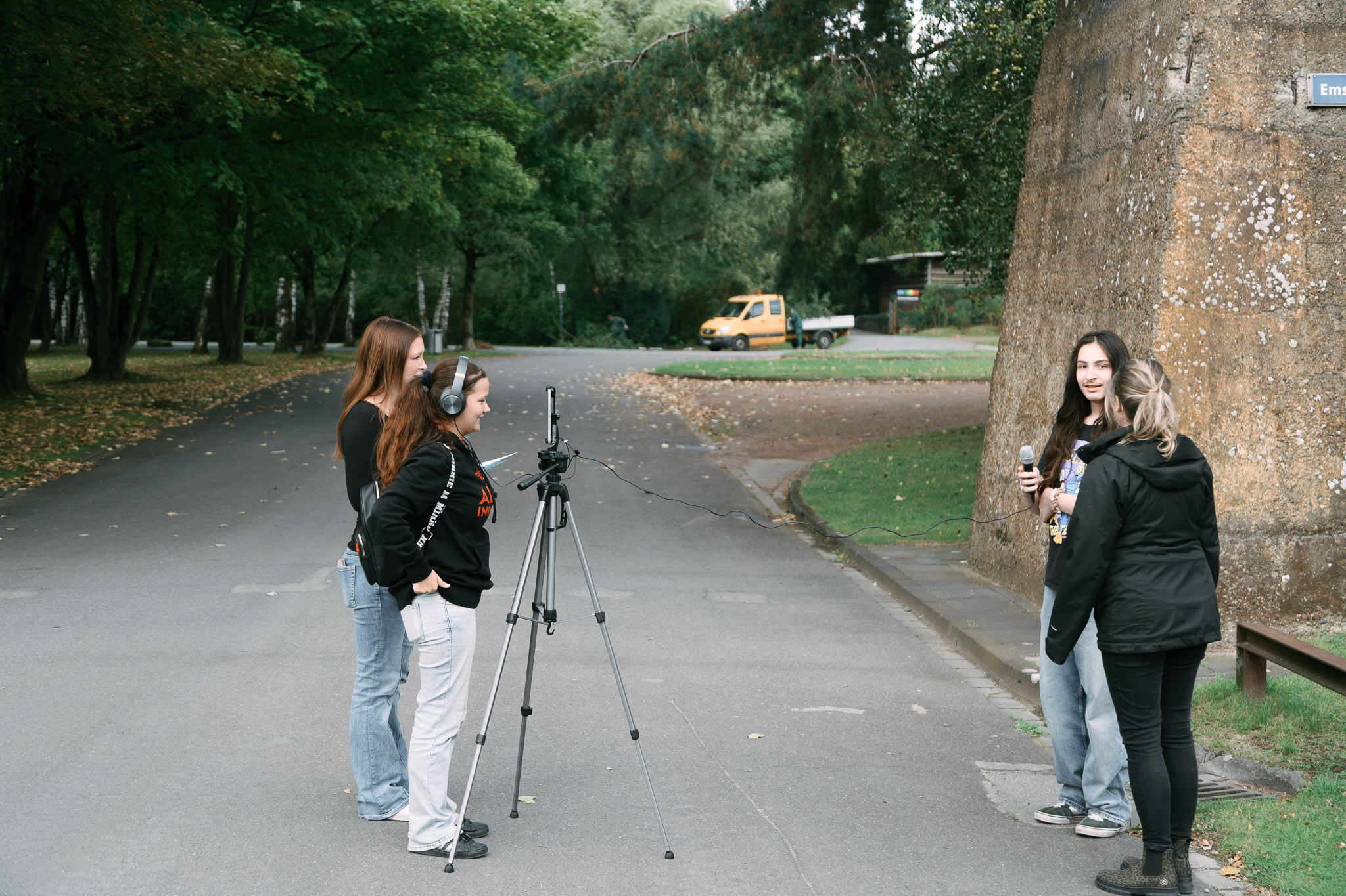 Drei junge Frauen führen ein Interview mit Mikrofon und Kamera auf einer Parkstraße.