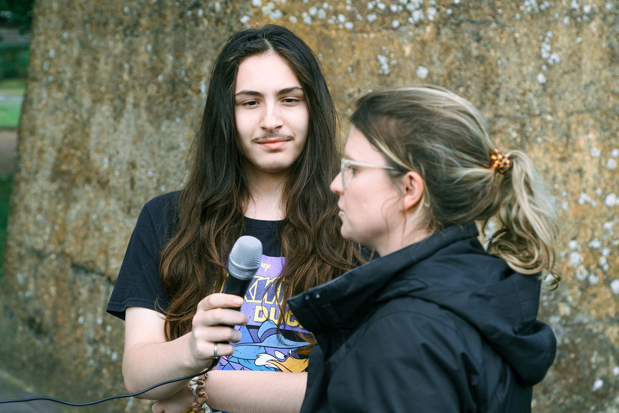 Zwei Personen stehen vor einer Steinwand, eine hält ein Mikrofon in der Hand.