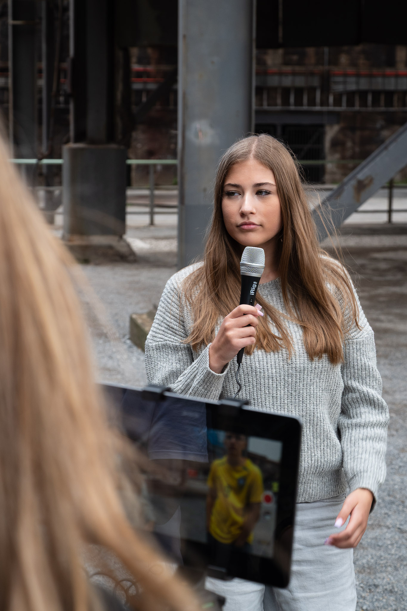 Junge Frau spricht mit Mikrofon vor Publikum in urbanem Außenraum mit Betonarchitektur.