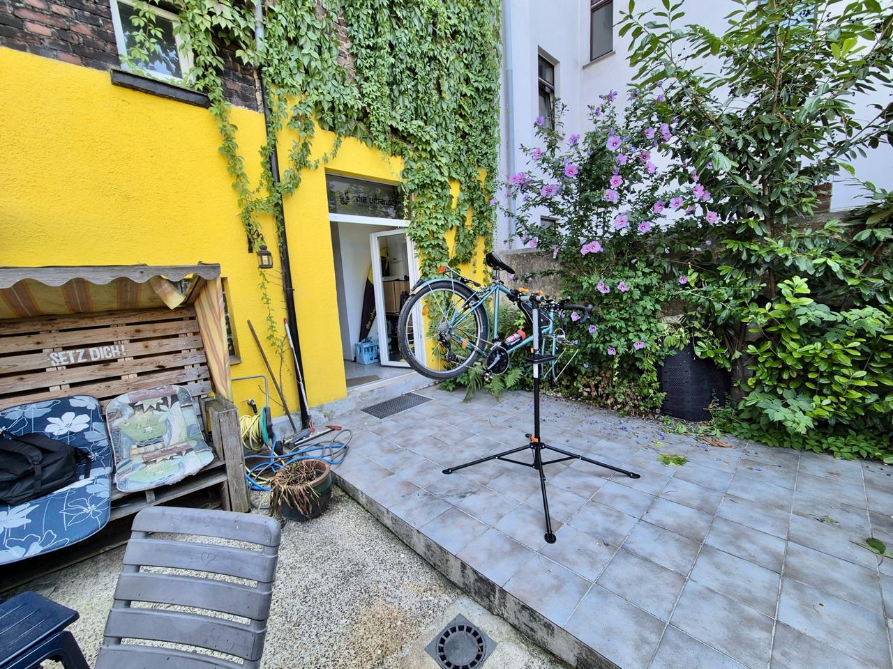 Innenhof mit gelbem Gebäude, Fahrrad auf Ständer, Kletterpflanzen und lila Blüten.