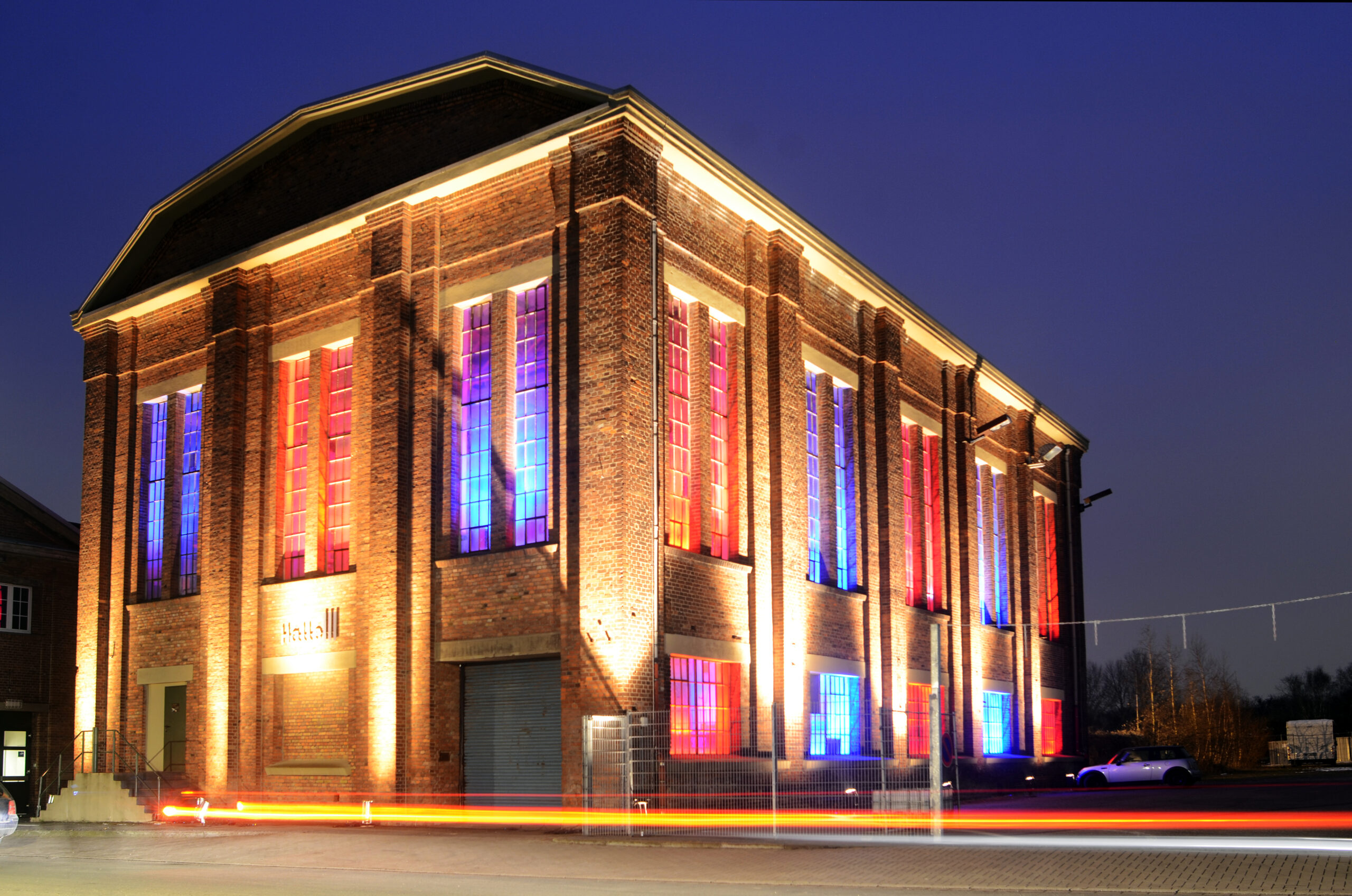 Beleuchtetes Backsteingebäude mit bunten Glasfenstern in Blau und Rot bei Dämmerung.
