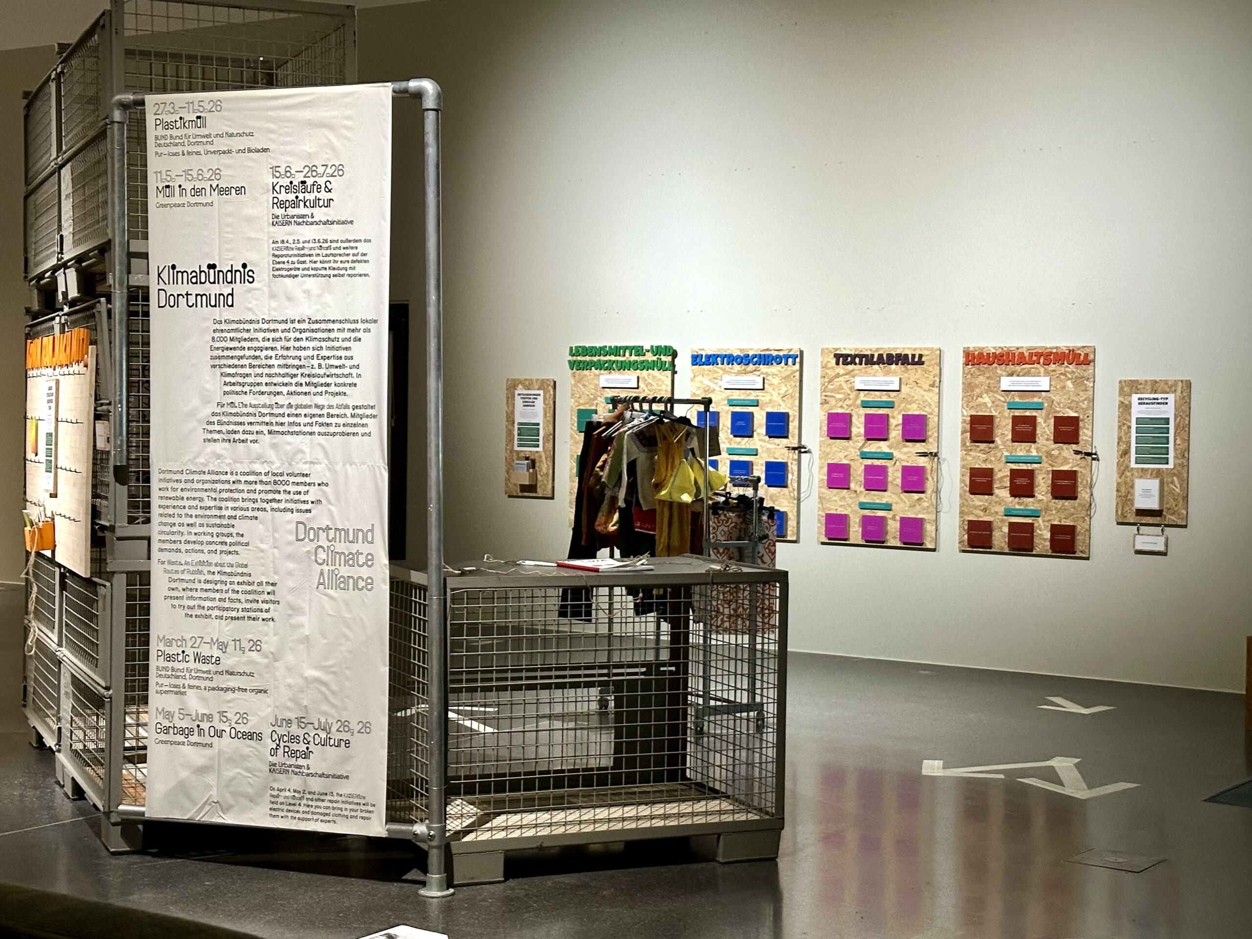 Ausstellungsaufbau mit Informationspanelen, Kleidungsständer und Materialmustern an der Wand.