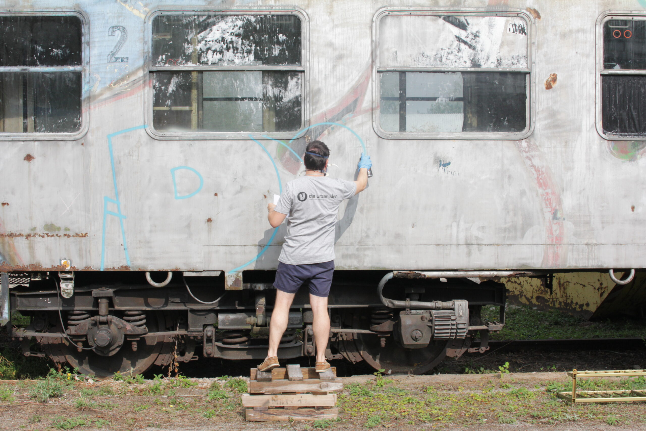 Ein Mann sprayt blau auf die Seite eines stillgelegten Eisenbahnwagens.