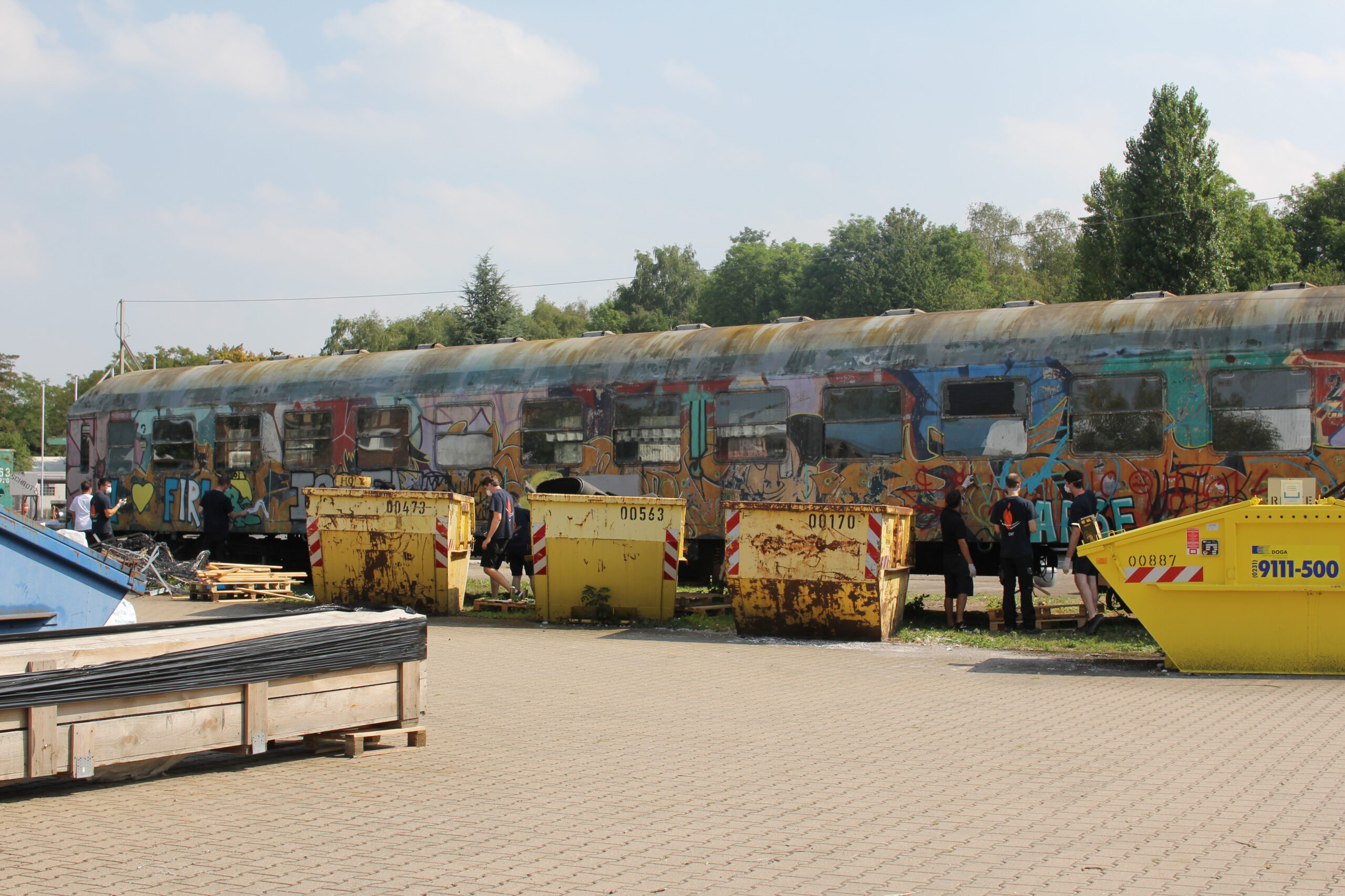 Bemalter Zug mit farbiger Graffiti-Kunst auf einem Gelände mit gelben Containern und Menschen.